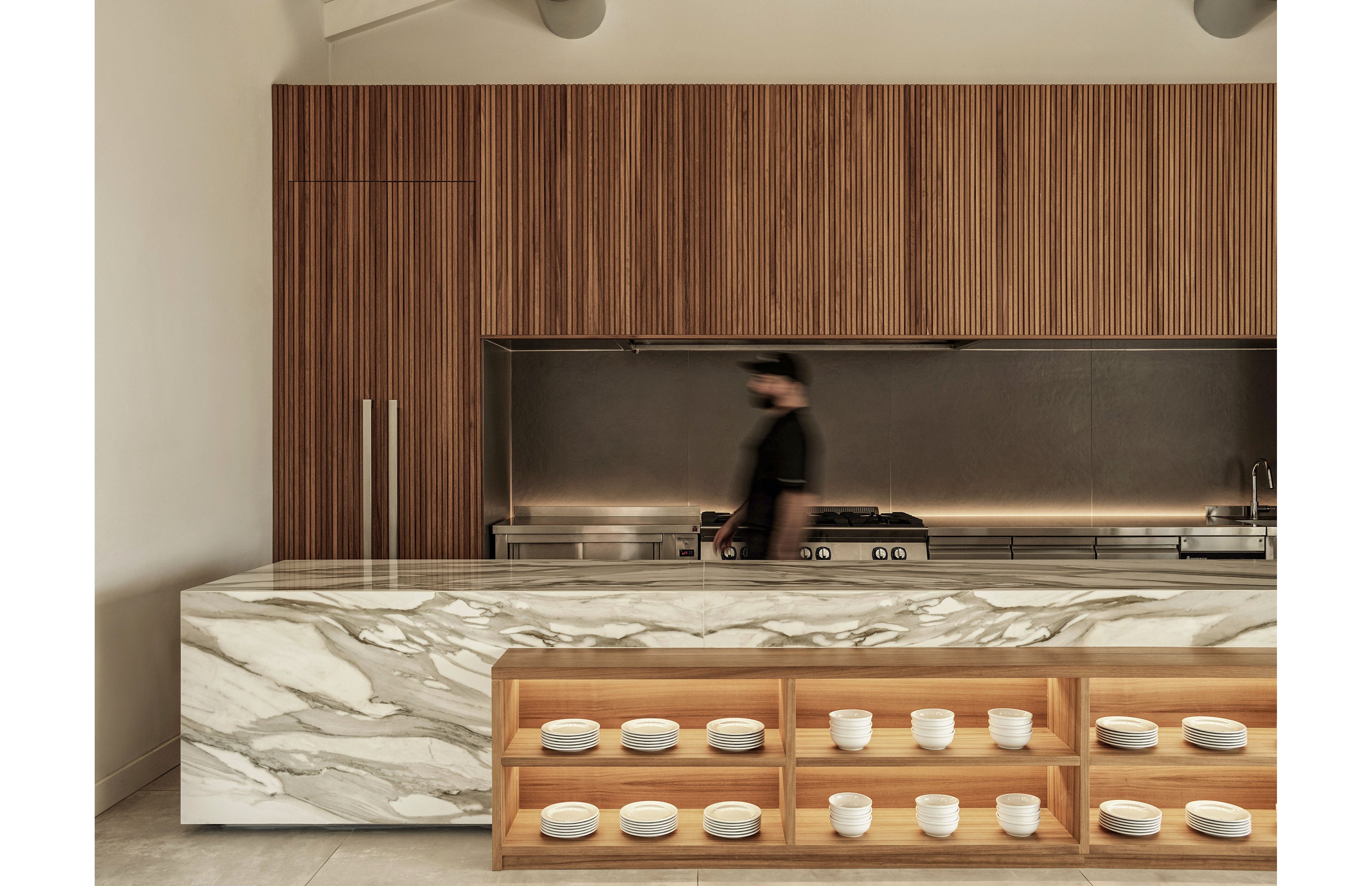 A contemporary kitchen features a large marble island with a built-in wooden shelf displaying neatly arranged white bowls. Vertical wood paneling covers the back wall, framing a sleek stainless steel cooking area. A blurred person in dark clothing walks past the stove, adding a sense of motion to the otherwise calm interior. Warm neutral tones and clean lines create a minimalist, sophisticated atmosphere.