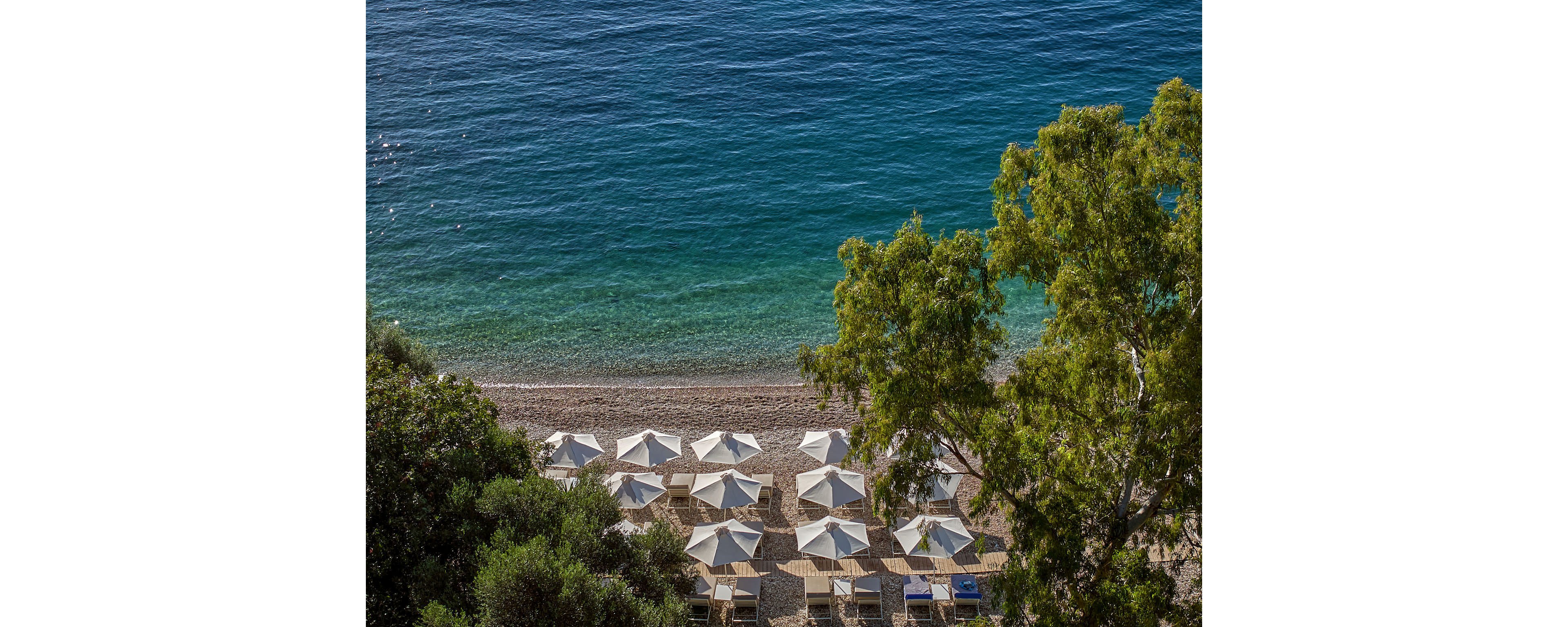 A tranquil beach resort is viewed from above, featuring neat rows of white umbrellas and lounge chairs on a sandy shoreline. Clear turquoise water gently meets the shore, creating a gradient of blue and green hues. Lush green trees frame the scene, adding natural shade and contrast to the bright coastal setting. The overall atmosphere is calm, inviting, and ideal for a relaxing seaside holiday.