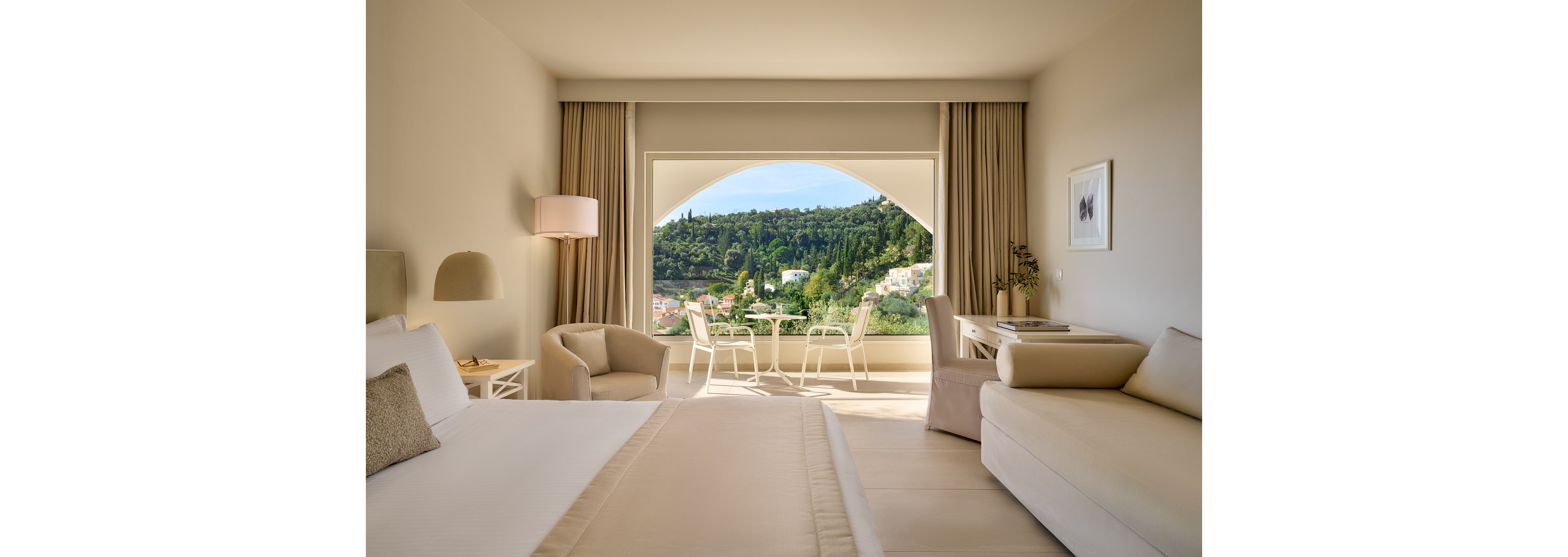 A spacious, modern hotel room features a neatly made bed and a light-colored sofa in a minimalist interior. Large curtains frame an arched opening that leads to a balcony with a table and chairs. Beyond the balcony, lush green hills and a small town are visible under clear daylight. Soft neutral tones and clean lines create a calm, inviting atmosphere ideal for relaxation.
