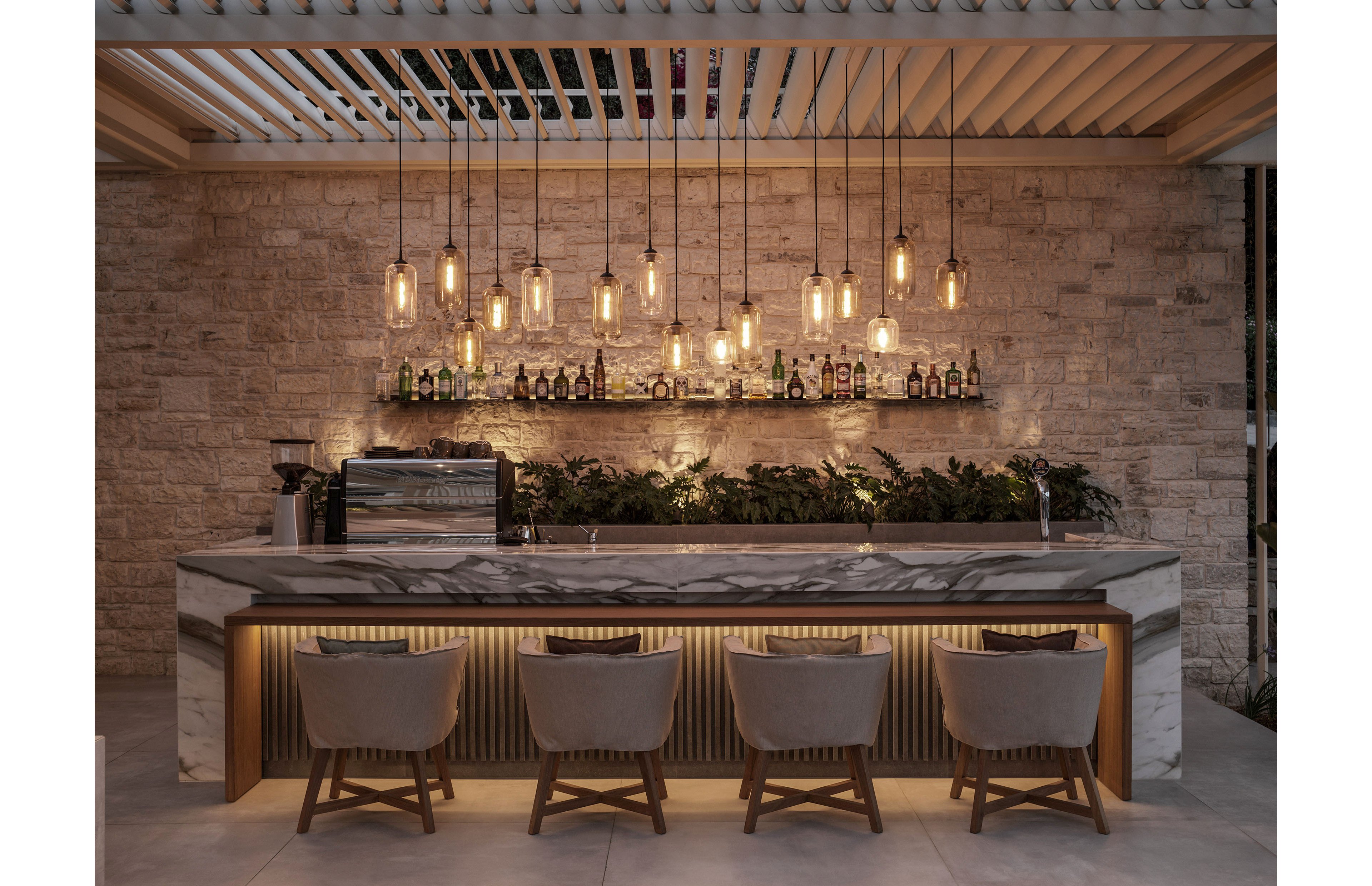 A contemporary bar interior features a long marble counter with four upholstered chairs neatly aligned in front. Warm pendant lights hang in a row above the bar, casting a soft glow over the stone wall backdrop. Bottles and glassware are arranged on a shelf, with greenery adding a natural accent beneath. The overall atmosphere is cozy and sophisticated, blending modern design with rustic textures.