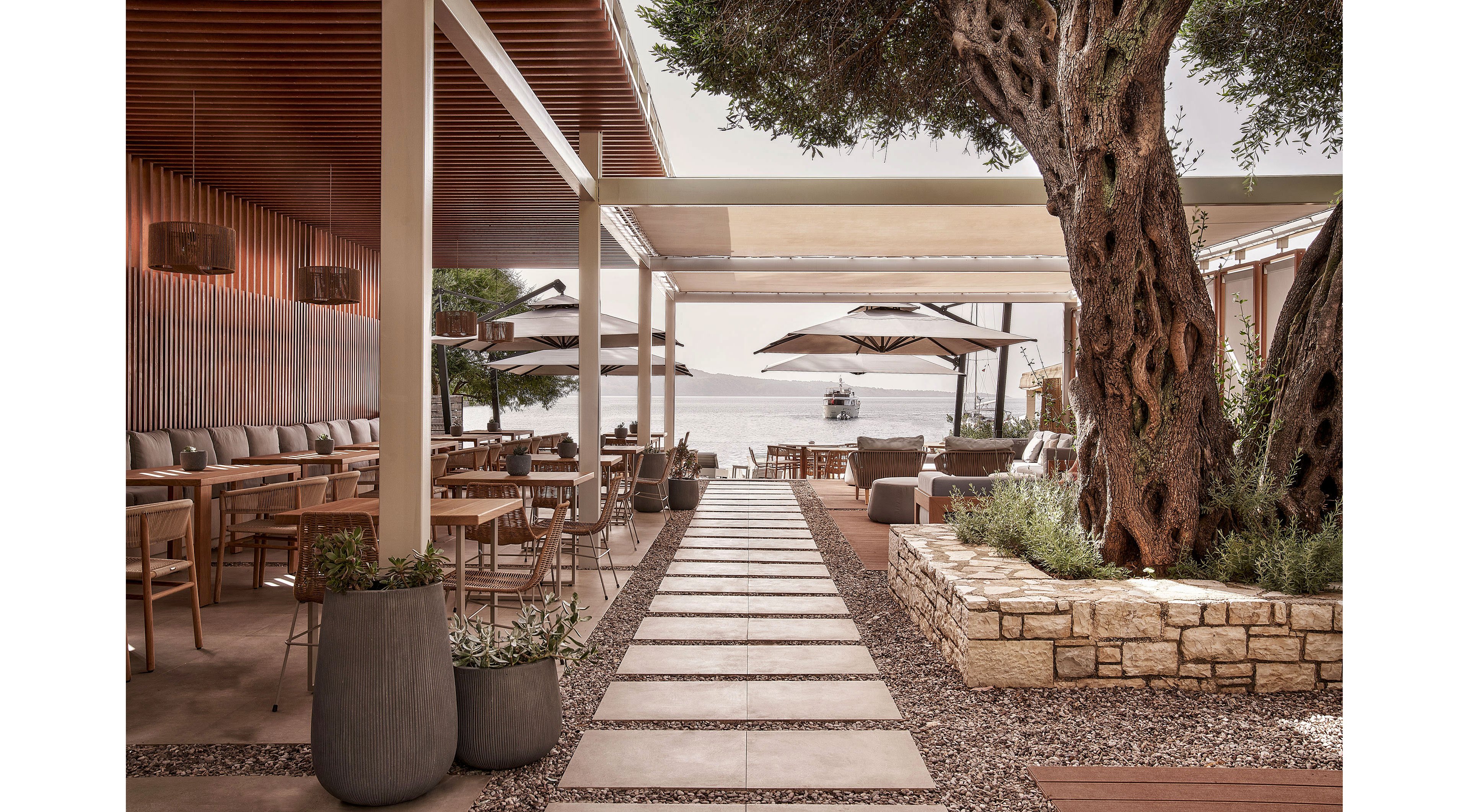 An elegant open-air restaurant terrace features wooden tables and cushioned seating arranged along a central stone walkway. Large potted plants and a mature tree add natural texture beside low stone walls and gravel landscaping. Neutral tones, wood slats, and fabric umbrellas create a warm, contemporary Mediterranean-inspired atmosphere. In the background, the sea and a small boat are visible, enhancing the relaxed coastal dining setting.
