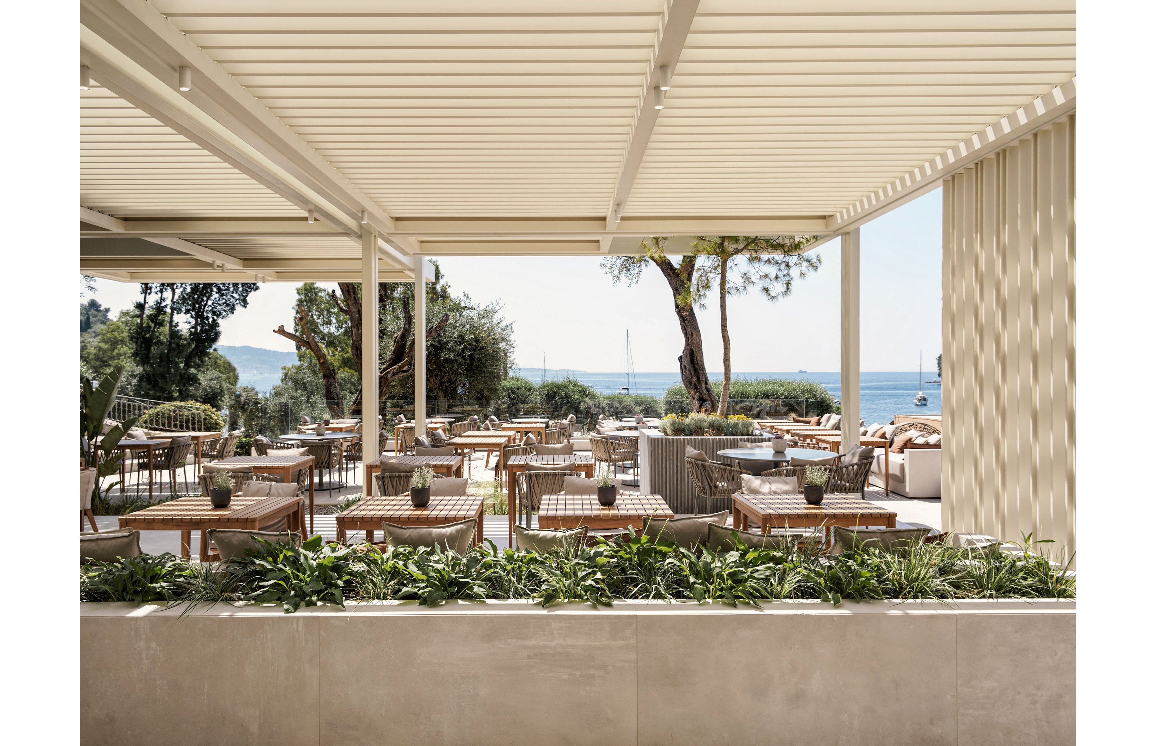 Contemporary outdoor restaurant seating is arranged beneath a sleek pergola overlooking the sea. Wooden tables and woven chairs are neatly aligned, with potted plants adding natural accents to the space. Lush greenery borders the terrace, leading the eye toward trees, water, and distant boats. The overall atmosphere is calm, airy, and inviting, ideal for relaxed coastal dining.