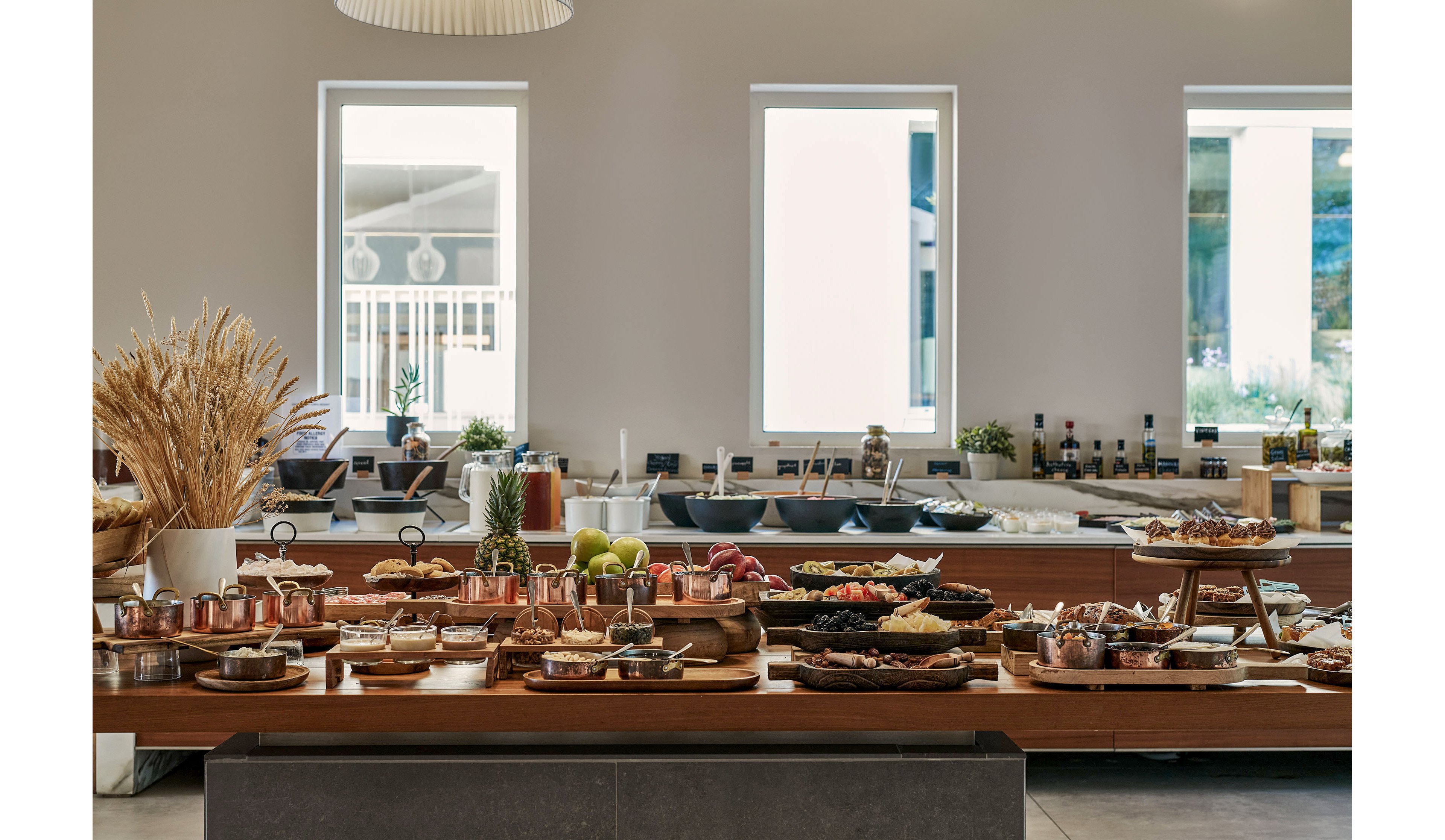 A long wooden buffet table is filled with an assortment of gourmet dishes and desserts in a modern dining space. Copper pots, wooden boards, and ceramic bowls display pastries, fruits, and savory items in an inviting arrangement. Large windows in the background let in natural light, revealing shelves with bottles and serving ware. Neutral tones and natural materials create a refined yet warm atmosphere suitable for upscale dining or events.