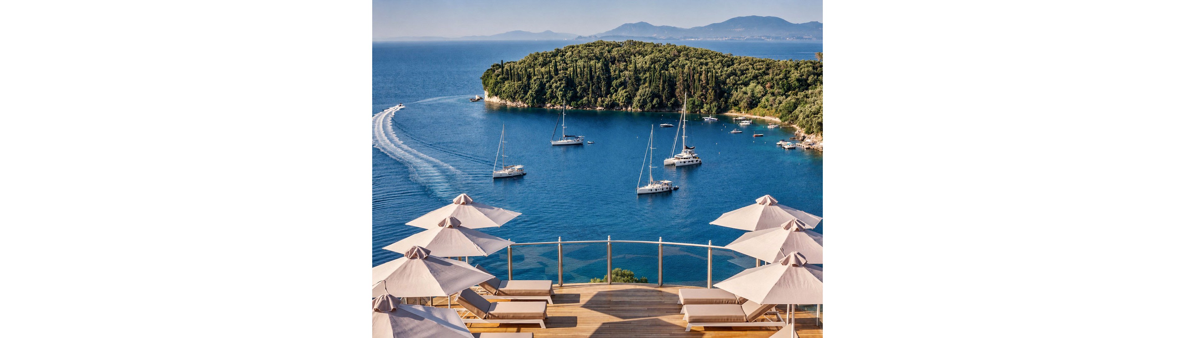 A luxurious waterfront terrace with sun loungers and umbrellas overlooks a calm blue bay. Several sailboats and yachts float on the water, leaving gentle wakes behind them. A lush green island rises in the middle distance, framed by hazy mountains on the horizon. The scene suggests a tranquil vacation setting with clear skies and inviting coastal views.