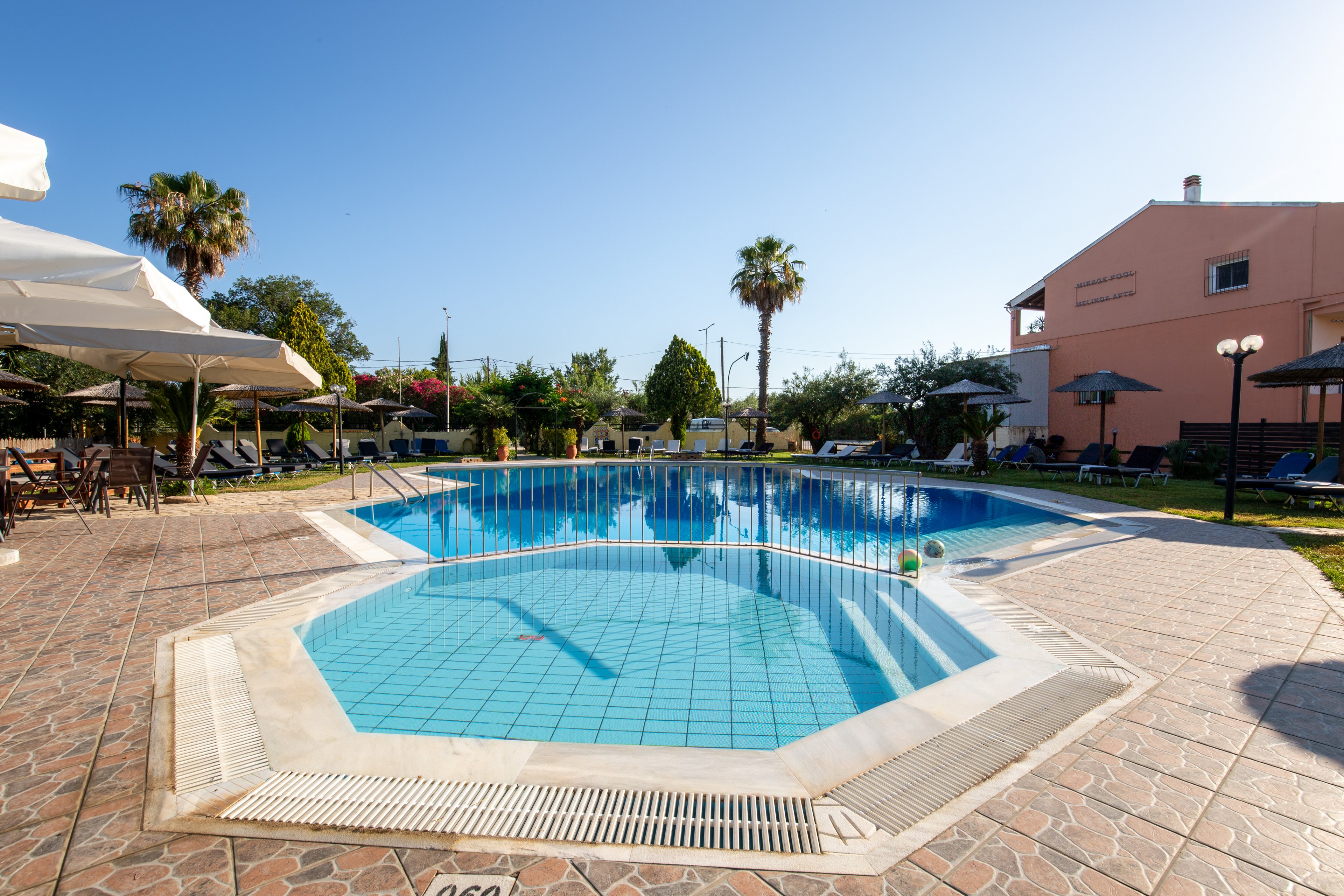 An outdoor resort swimming pool is shown under a clear blue sky, with a smaller hexagonal pool in the foreground. Sun loungers and umbrellas line the poolside area, suggesting a relaxing vacation setting. Palm trees and landscaped greenery surround the pool, while a light-colored building stands in the background. The bright sunlight and reflections on the water create a warm, inviting atmosphere.