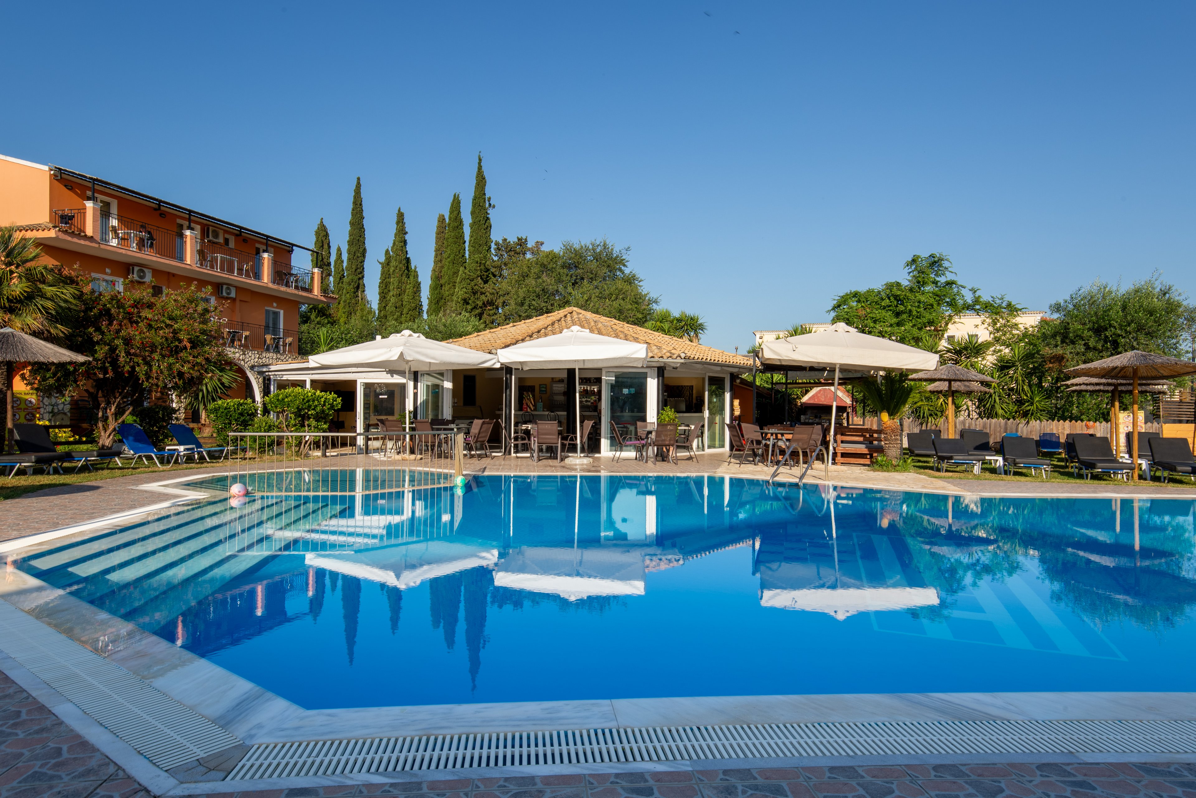 A large outdoor swimming pool reflects the clear blue sky and surrounding buildings at a resort. Sun loungers and parasols line the poolside, creating a relaxed vacation atmosphere. A central pool bar or café with seating is visible, framed by tall trees and greenery. The scene suggests a warm, sunny day ideal for leisure and holiday travel.
