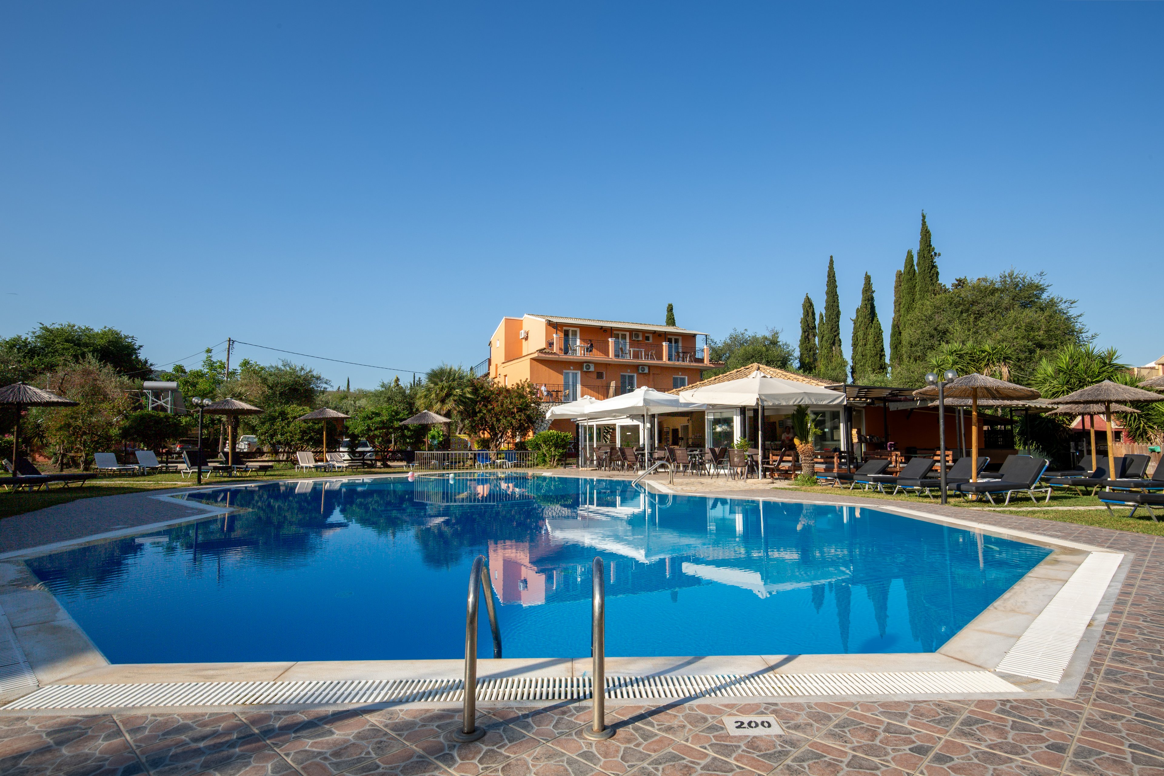 A large outdoor swimming pool reflects the clear blue sky at a resort. Sun loungers and straw parasols surround the pool, with guests relaxing in the shade. In the background, an orange hotel building and tall cypress trees frame the scene. The atmosphere is calm and inviting, suggesting a warm vacation day by the water.