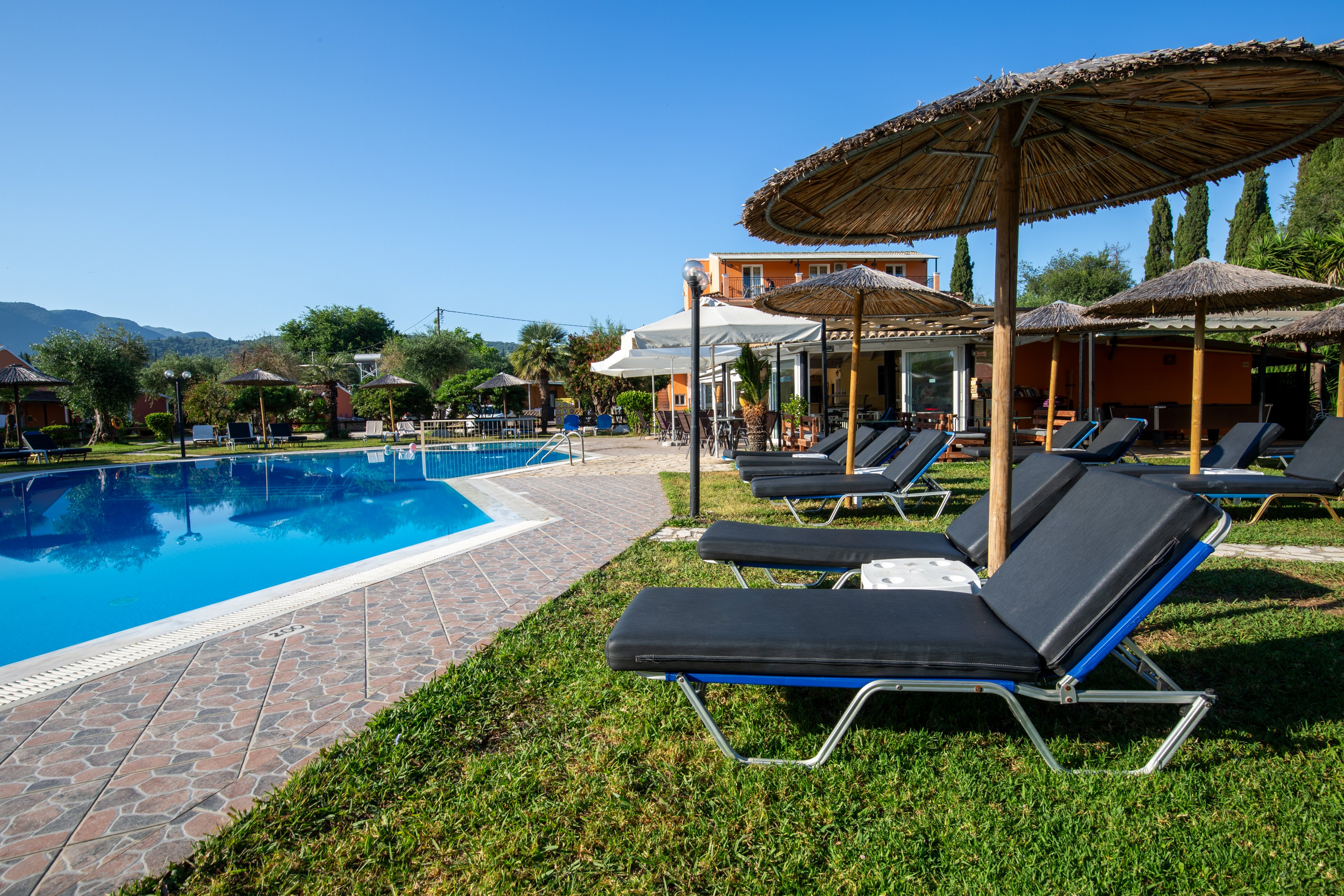 A row of empty sun loungers sits on a grassy area beside a clear blue swimming pool. Straw parasols provide shade along the tiled poolside path, with resort buildings visible in the background. The scene is set on a bright sunny day with a clear sky and surrounding greenery. The tranquil atmosphere suggests a relaxing vacation destination.