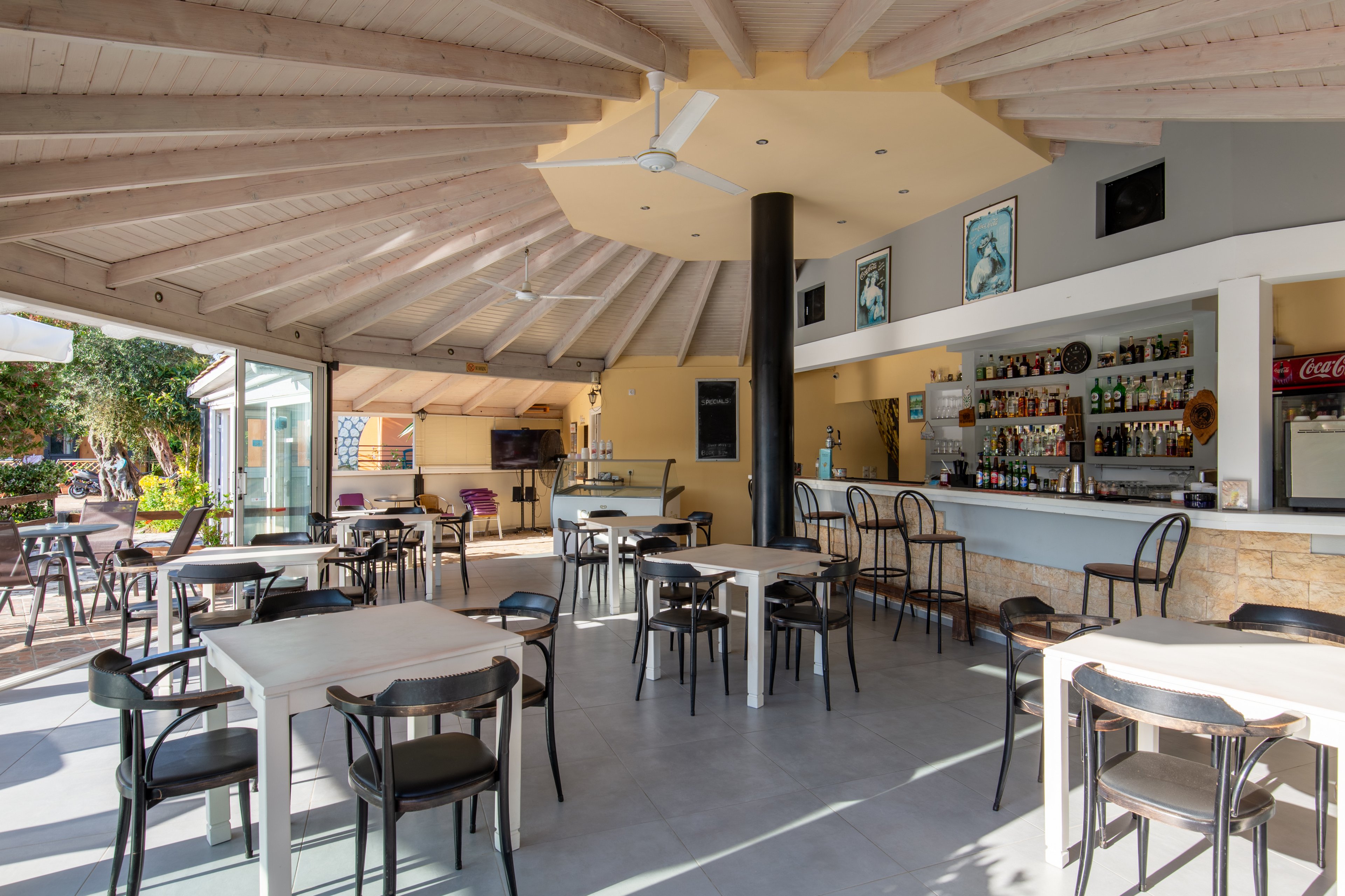 An open-air bar and café interior features multiple tables and chairs arranged under a wooden, vaulted ceiling with a central column and ceiling fan. The bar counter on the right displays bottles, glassware, and a coffee machine, with high stools lined along the front. Large openings connect the space to an outdoor garden area, allowing natural light to fill the room. The overall atmosphere is casual and inviting, suitable for relaxed dining or drinks at a resort or holiday venue.