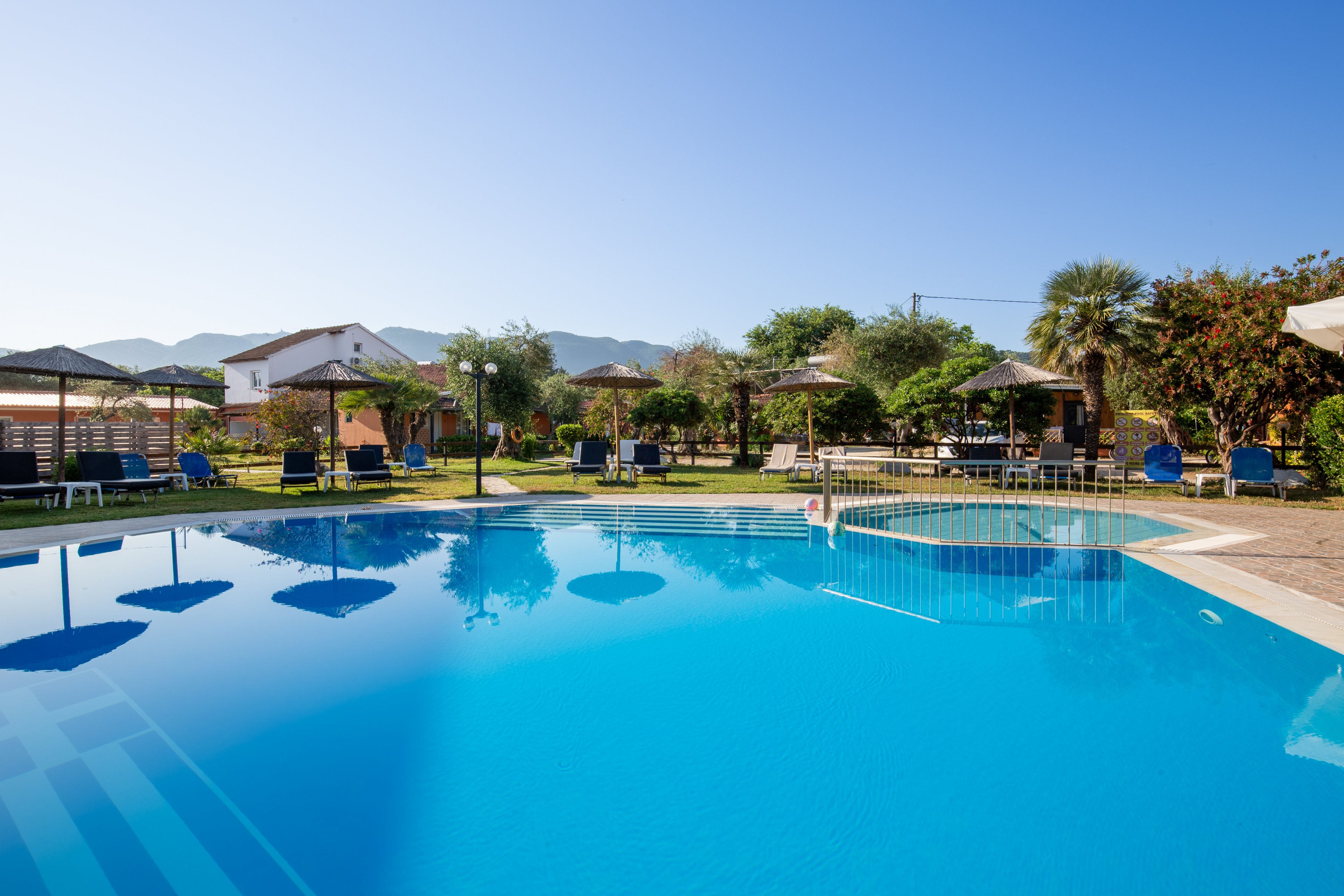 A clear blue outdoor swimming pool is shown in a sunny resort setting. Sun loungers and straw parasols surround the pool, with landscaped greenery and palm trees adding a tropical feel. Residential-style buildings and distant mountains are visible in the background under a bright blue sky. The calm water and empty seating create a peaceful vacation atmosphere.