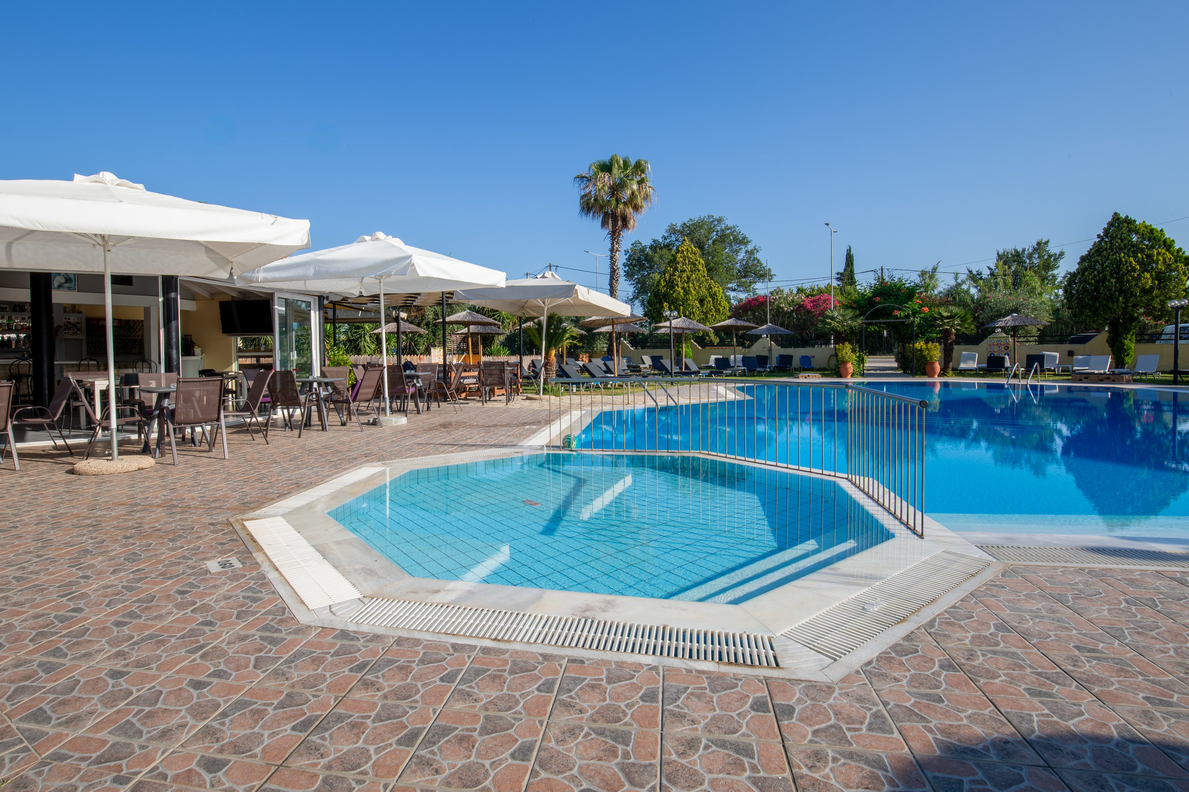 An outdoor resort swimming pool is shown under a clear blue sky, featuring a smaller octagonal pool section in the foreground. The surrounding patio is paved with stone tiles and lined with tables, chairs, and large white umbrellas. In the background, lush green trees, flowering shrubs, and sun loungers suggest a relaxing holiday setting. The overall scene conveys a bright, tranquil atmosphere ideal for leisure and relaxation.
