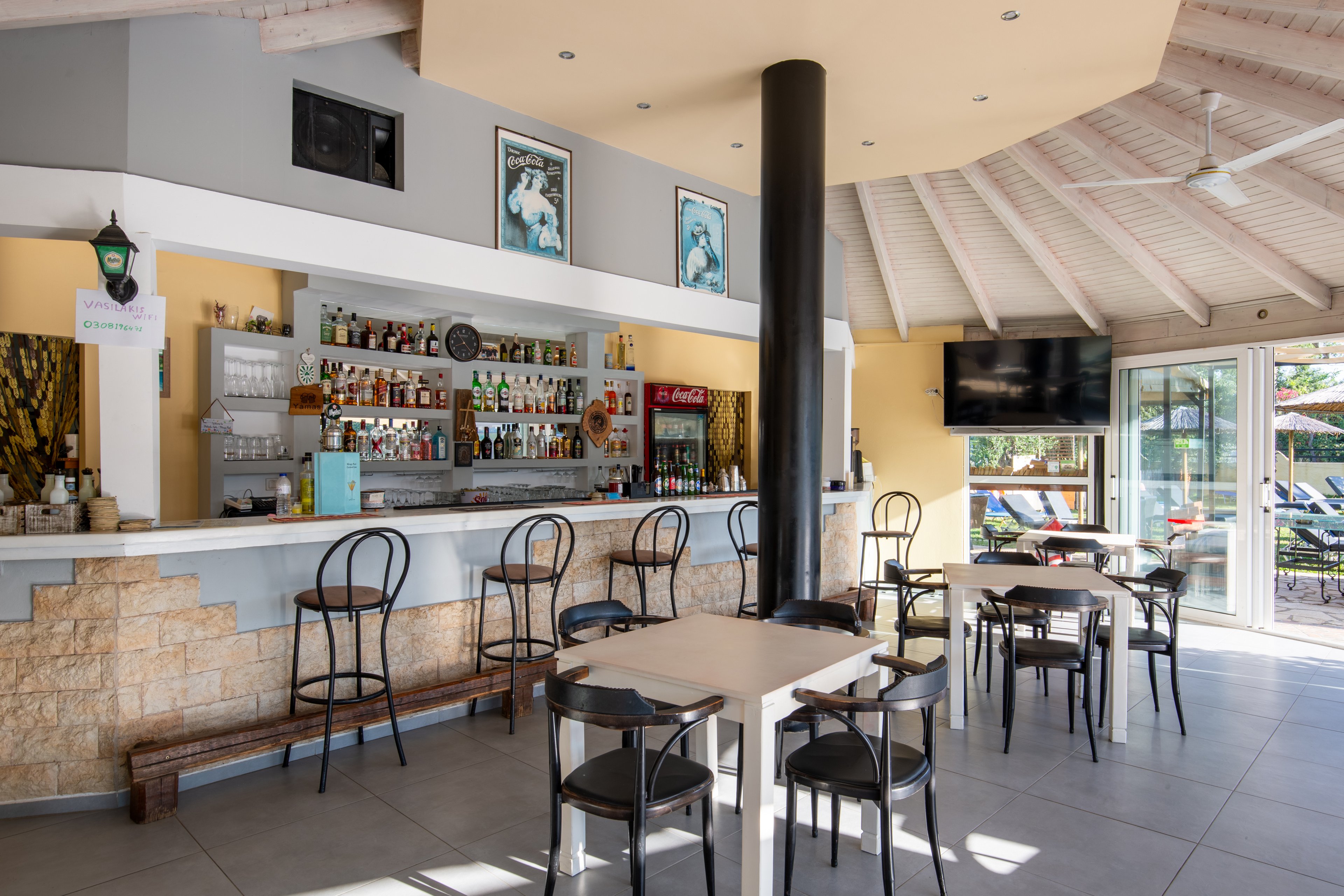 A sunlit bar interior features a long counter lined with bottles and glassware. Several black metal barstools and square tables with chairs are arranged across the tiled floor. Large windows and glass doors open to an outdoor poolside area, while a flat-screen television is mounted on the wall. The space has a light, airy feel with neutral colors and a high, white-beamed ceiling.