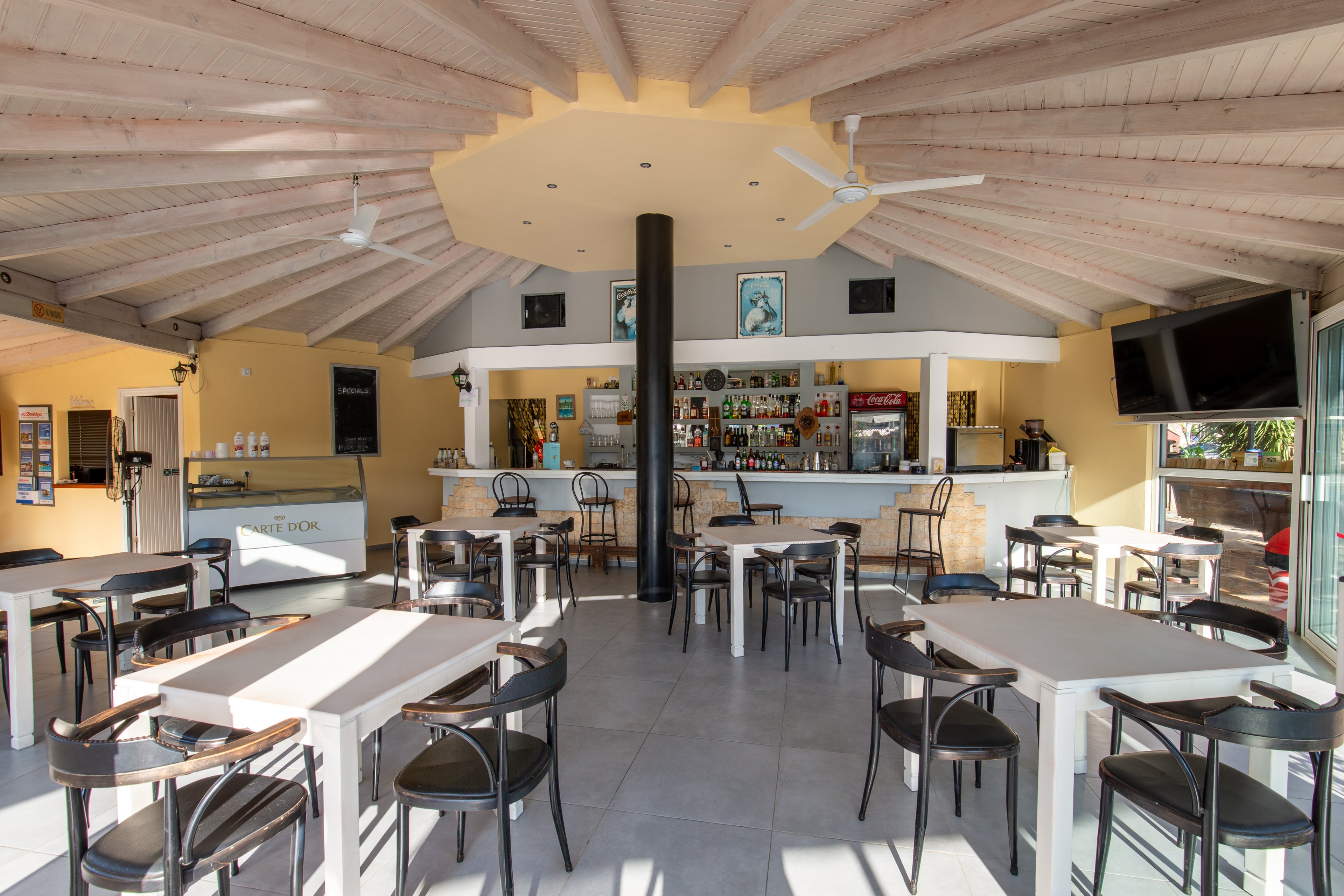 Spacious cafe bar interior with multiple tables and chairs arranged neatly. The setting features a high wooden ceiling with exposed beams and ceiling fans, creating an airy atmosphere. The bar counter at the back displays bottles, glasses, and a coffee machine, with framed artwork above. Large windows and doors allow natural light to fill the room, highlighting the clean, modern decor. No people are present, emphasizing the calm and orderly environment.