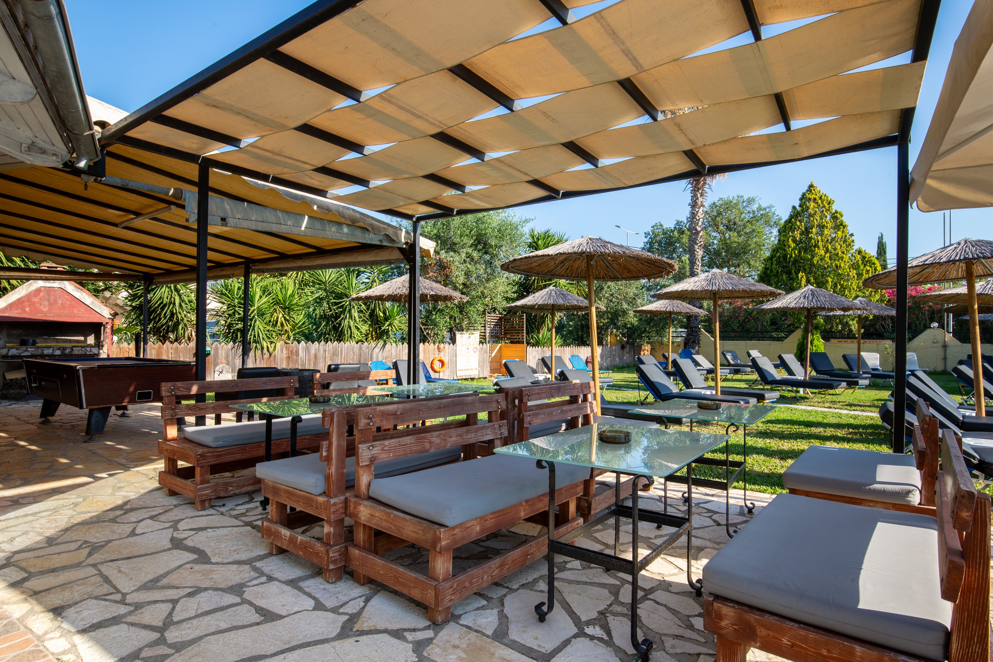 An outdoor resort lounge area features wooden benches with gray cushions arranged around glass-topped tables under a large pergola. The stone-paved patio opens onto a grassy lawn lined with reclining sunbeds and straw parasols. In the background, lush greenery and trees create a relaxed, tropical atmosphere. The scene is brightly lit, suggesting a sunny day ideal for leisure and relaxation.
