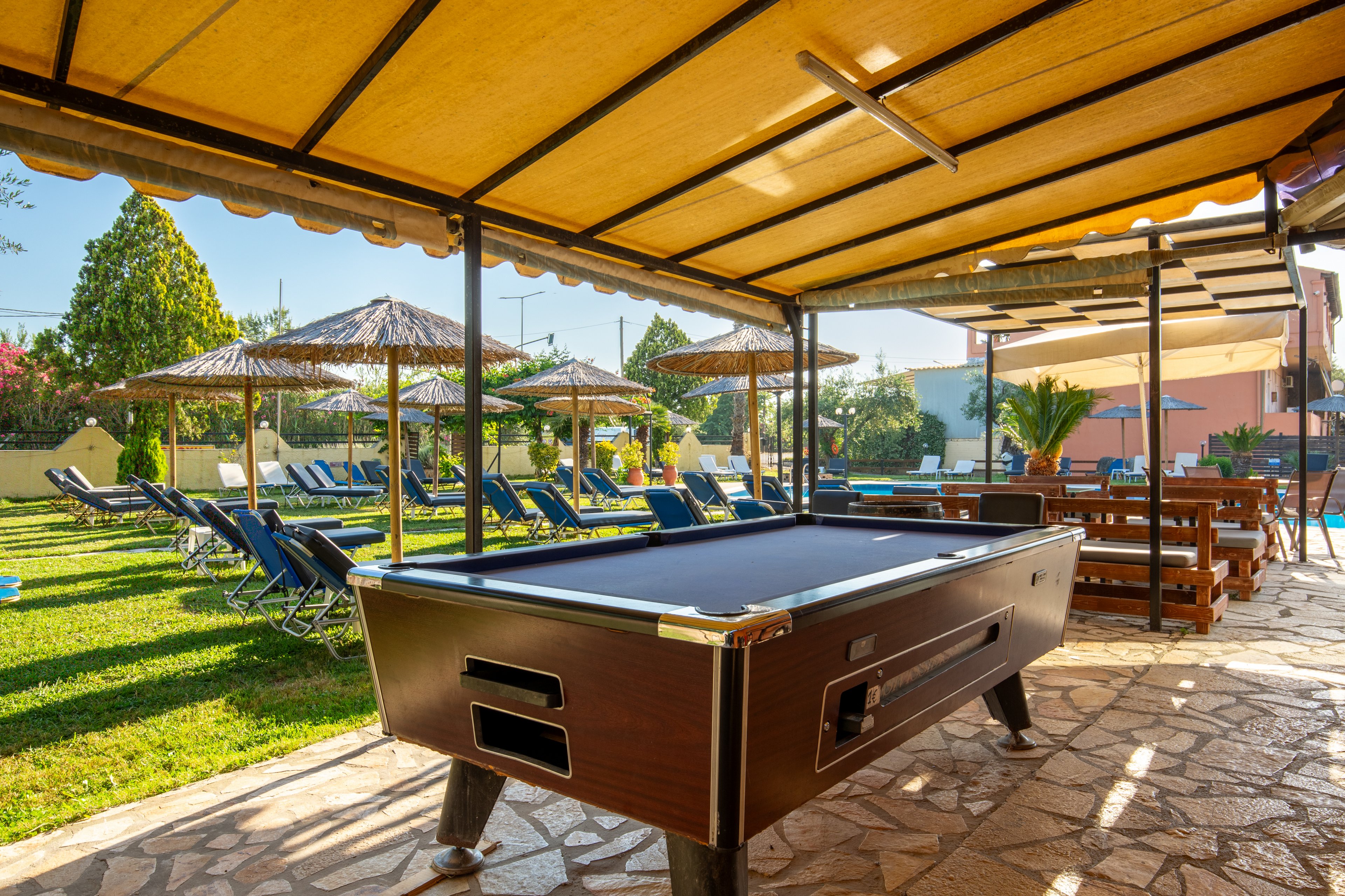 A wooden pool table stands under a shaded canopy in an outdoor leisure area. Behind it, rows of blue sun loungers line a grassy garden with straw parasols. The setting appears to be a resort or hotel yard, with warm sunlight and clear skies creating a relaxed vacation atmosphere. Stone paving, green plants, and nearby buildings complete the inviting recreational scene.