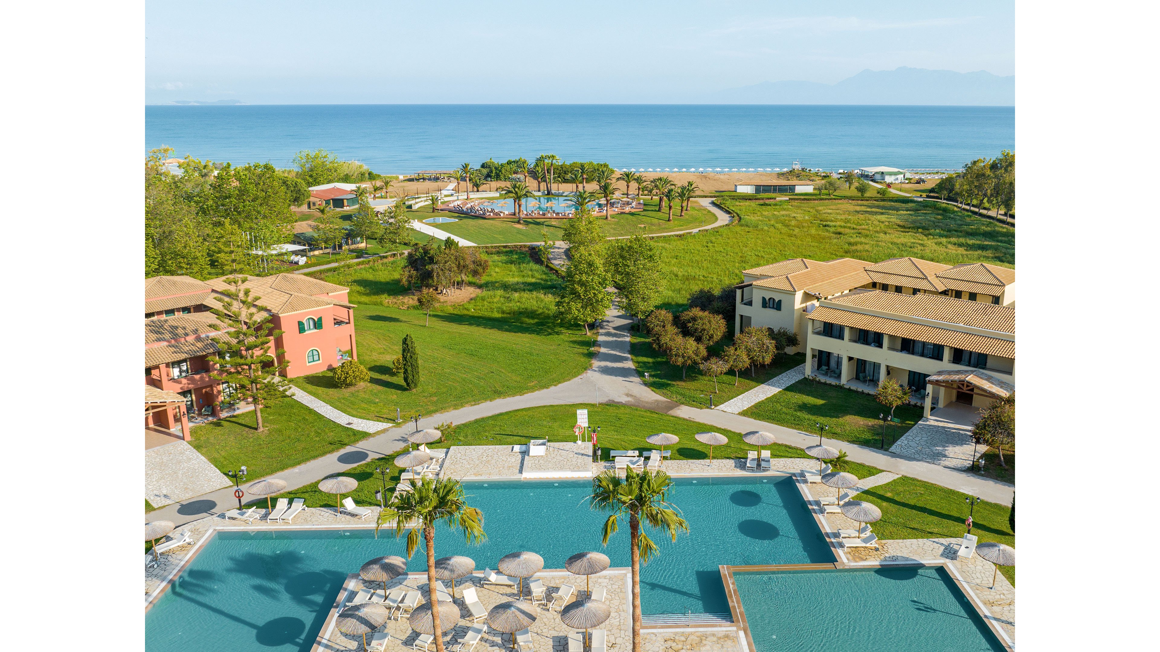 Aerial view of a seaside resort featuring multiple outdoor swimming pools in the foreground. Sun loungers and umbrellas are arranged neatly around the pools, with landscaped lawns and pathways connecting the buildings. Low-rise villas with terracotta roofs sit among trees and greenery, leading toward a distant pool and palm trees near the shoreline. The calm blue sea stretches to the horizon, with mountains faintly visible in the background, creating a tranquil vacation atmosphere.