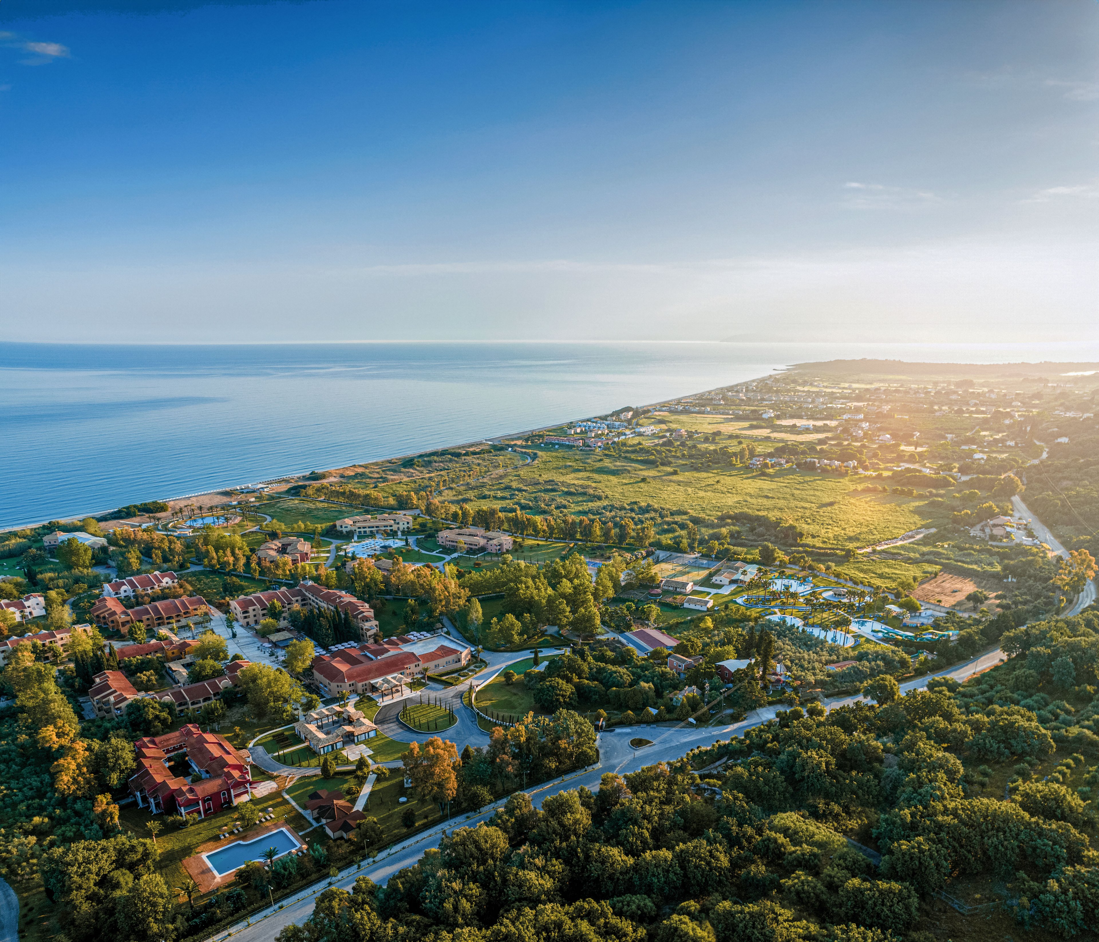 Aerial view of a coastal resort area stretches along the shoreline under a clear blue sky. Residential villas, pools, and landscaped gardens are surrounded by lush greenery and farmland. The calm sea forms a wide horizon, while the warm golden sunlight bathes the scene, creating long shadows and a tranquil atmosphere. Roads and scattered buildings indicate a peaceful vacation destination near the water.