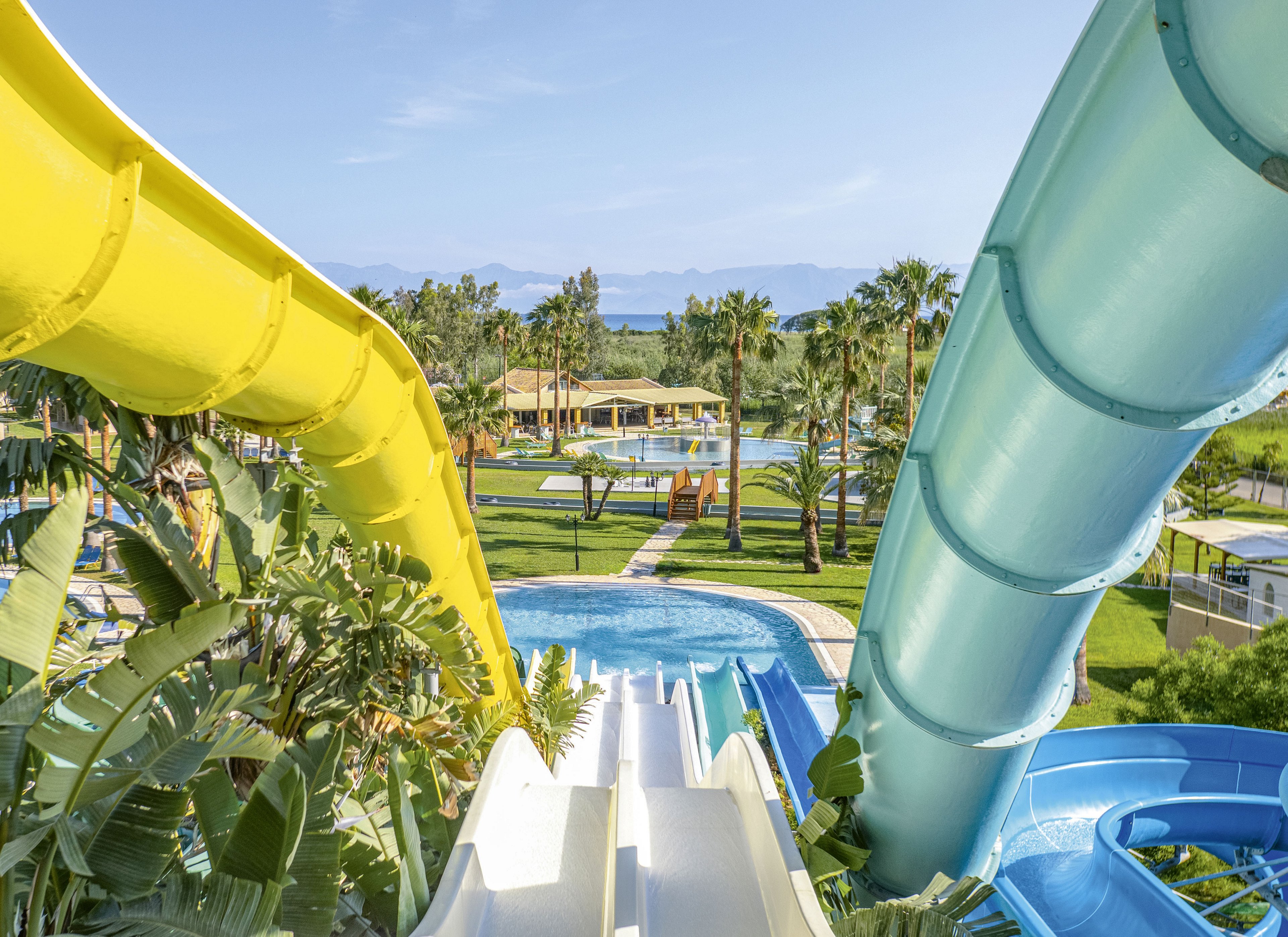 Bright yellow, white, and blue water slides curve down toward a clear blue pool in a lush resort setting. Palm trees, green lawns, and sunlit pathways surround the aquatic area, creating a vibrant vacation atmosphere. In the distance, low buildings and shaded structures sit beneath a clear sky with faint mountains on the horizon. The overall scene suggests a fun, family-friendly water park in a warm, tropical climate.