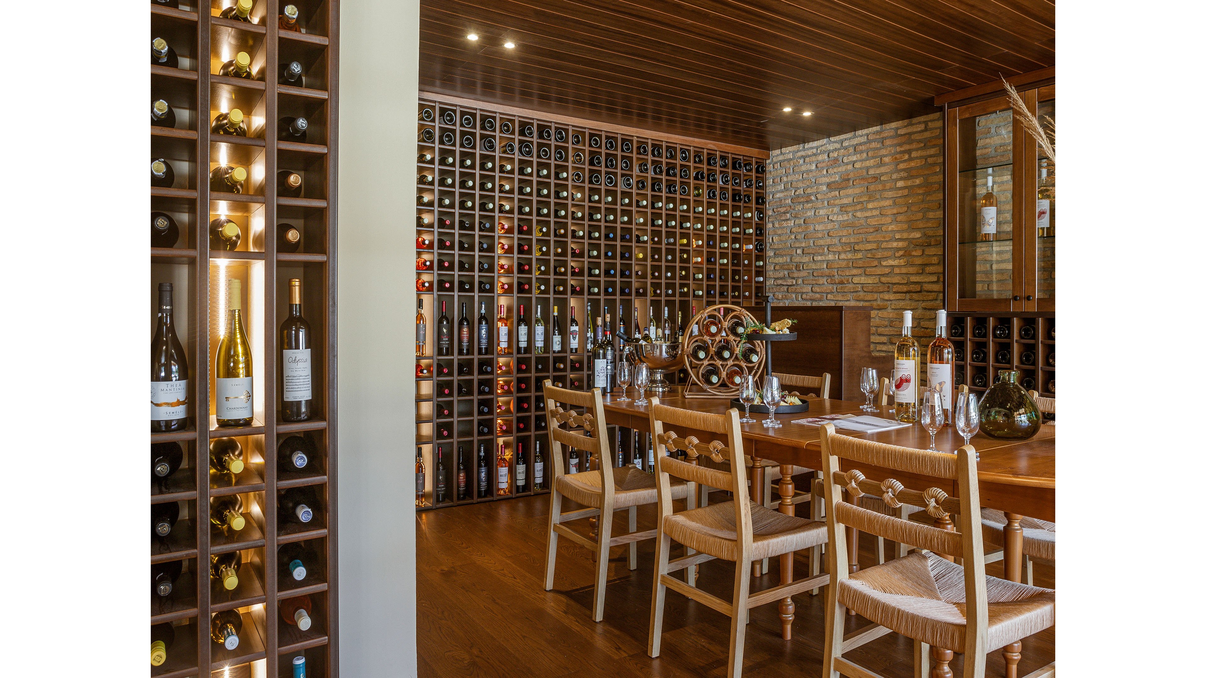Warm wooden wine cellar featuring a large collection of bottles neatly stored in wall racks. A wooden table with chairs is set for a wine tasting, with several bottles and glasses arranged on its surface. Brick walls and wood panel ceiling create an intimate, rustic atmosphere. Soft lighting highlights the wine displays and enhances the inviting, elegant setting.