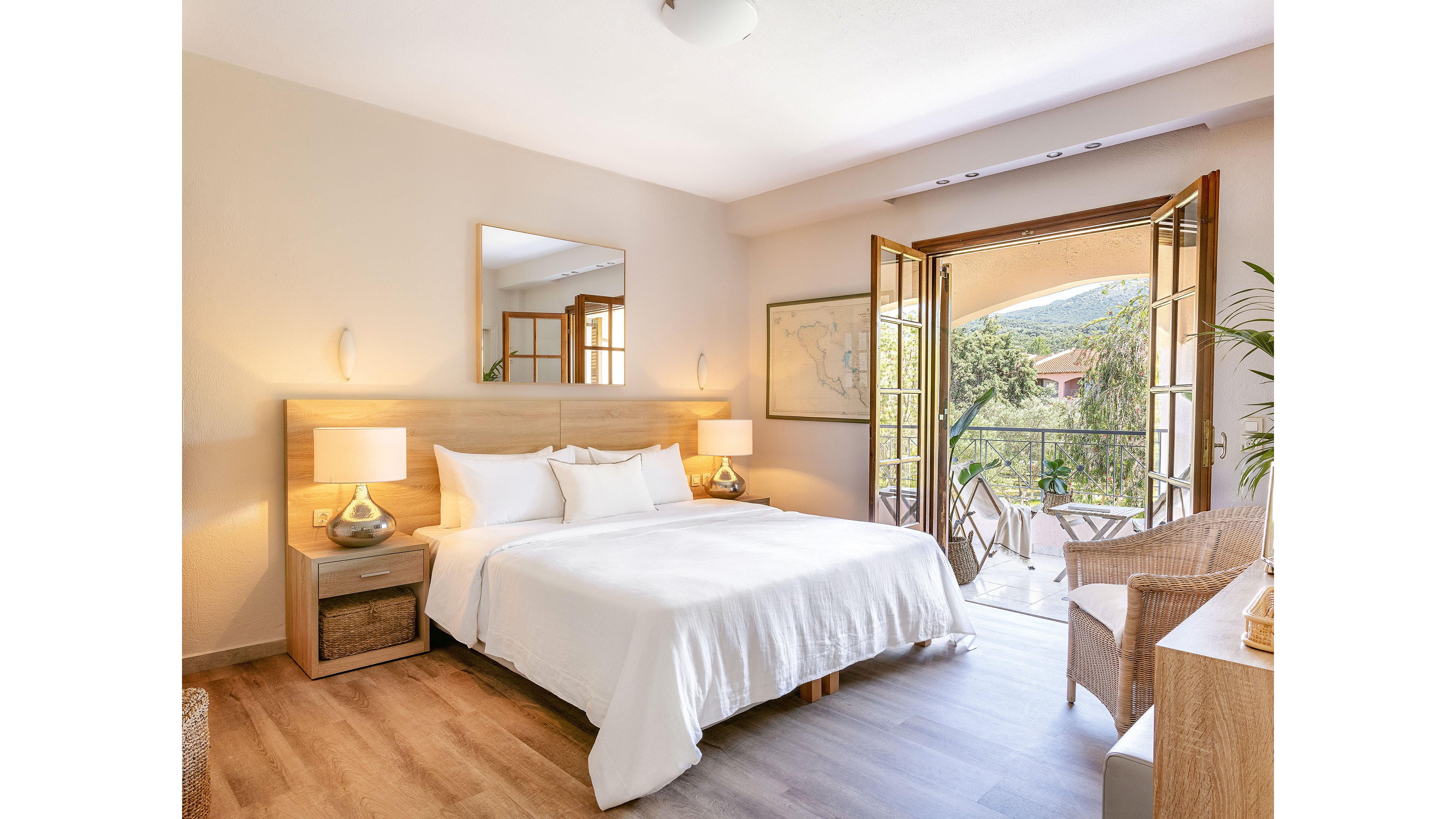 A neatly made double bed with white linens sits in a bright, modern bedroom. Matching bedside tables with lamps flank the bed, and a large mirror hangs above the headboard. French doors open to a balcony with outdoor chairs and a table, overlooking greenery and distant hills. Soft neutral tones and natural light create a calm, inviting atmosphere.