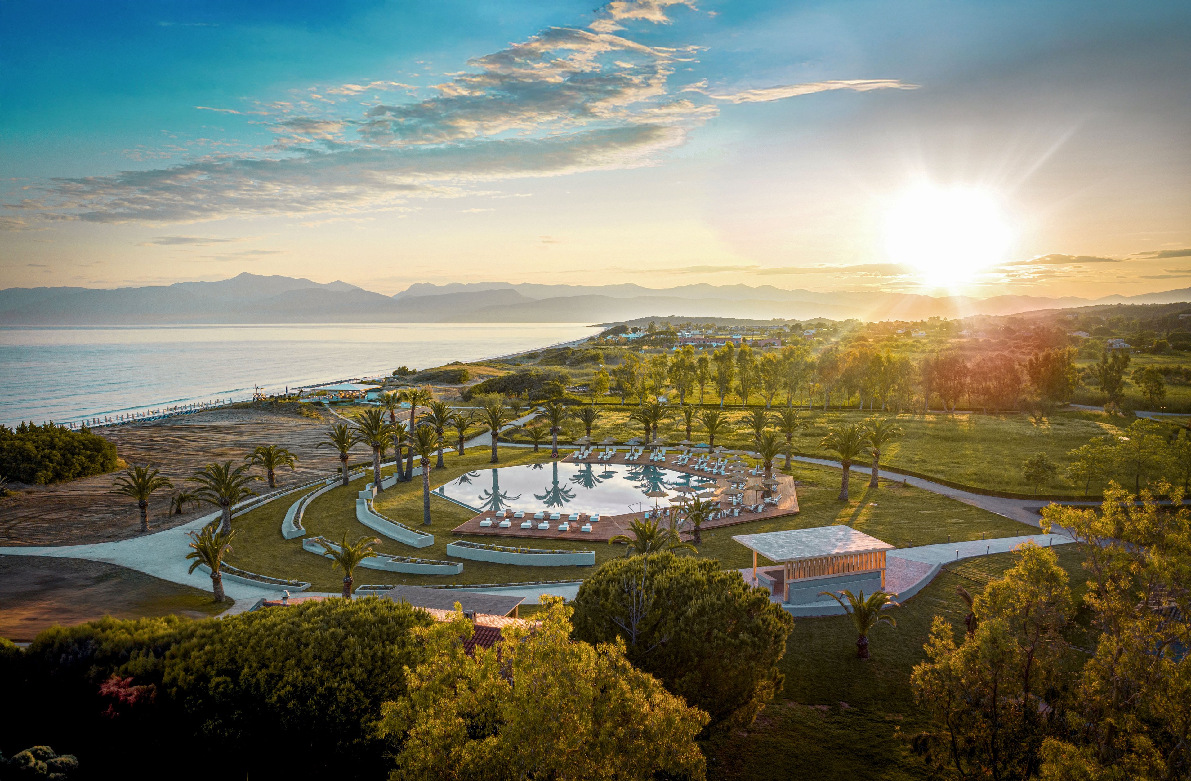 A large circular swimming pool is surrounded by palm trees in a landscaped coastal resort. The scene overlooks a calm sea with distant mountains under a bright setting or rising sun. Curved walkways, green lawns, and scattered trees frame the recreational area. The warm sunlight creates long shadows and a serene, inviting atmosphere.