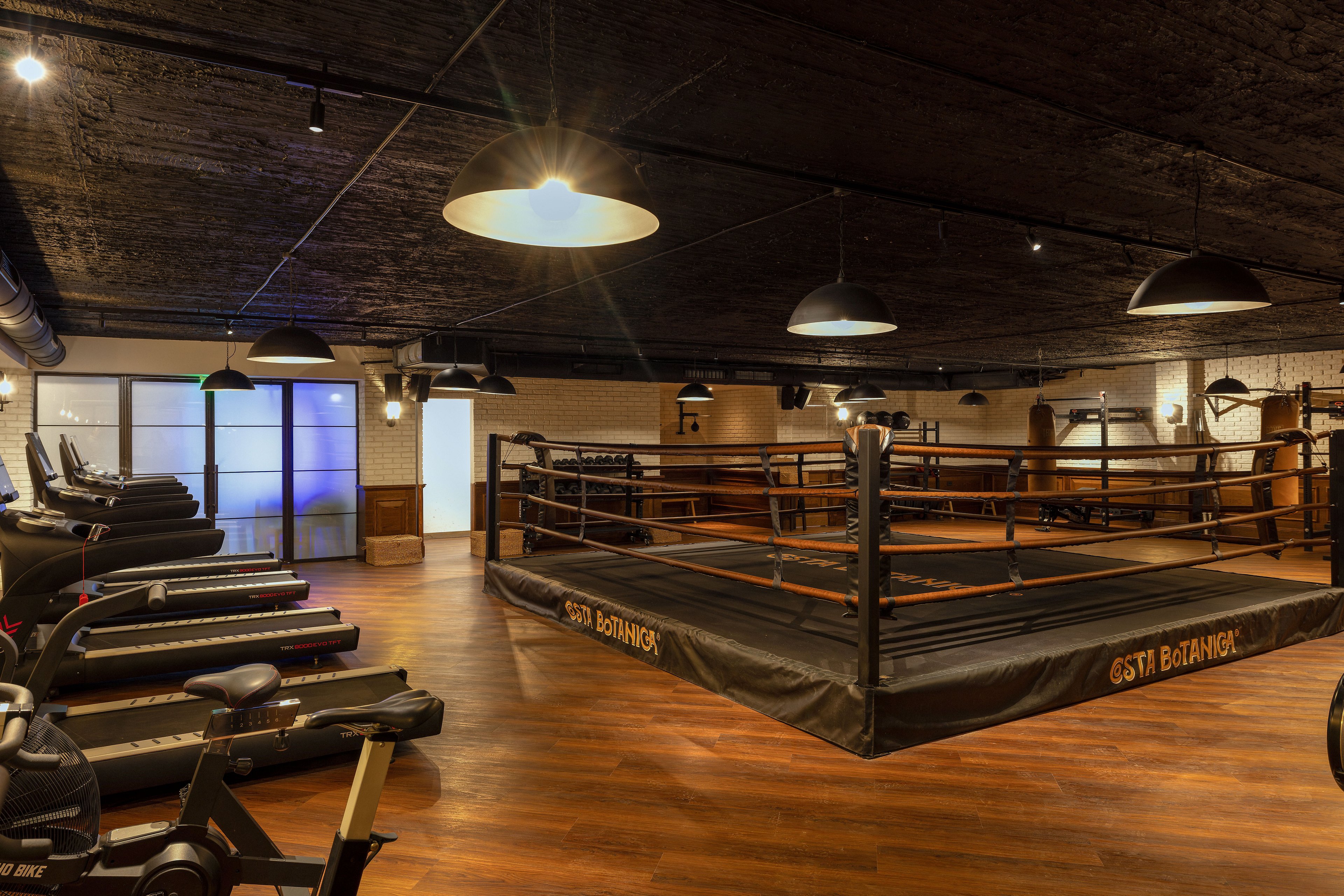 A spacious indoor boxing gym features a central boxing ring surrounded by wooden flooring. Cardio equipment, including treadmills, lines one side of the room, while industrial-style lighting hangs from the dark ceiling. The setting combines warm ambient light with contemporary design elements, creating a professional yet inviting training environment. No people are present, emphasizing the equipment and space itself.