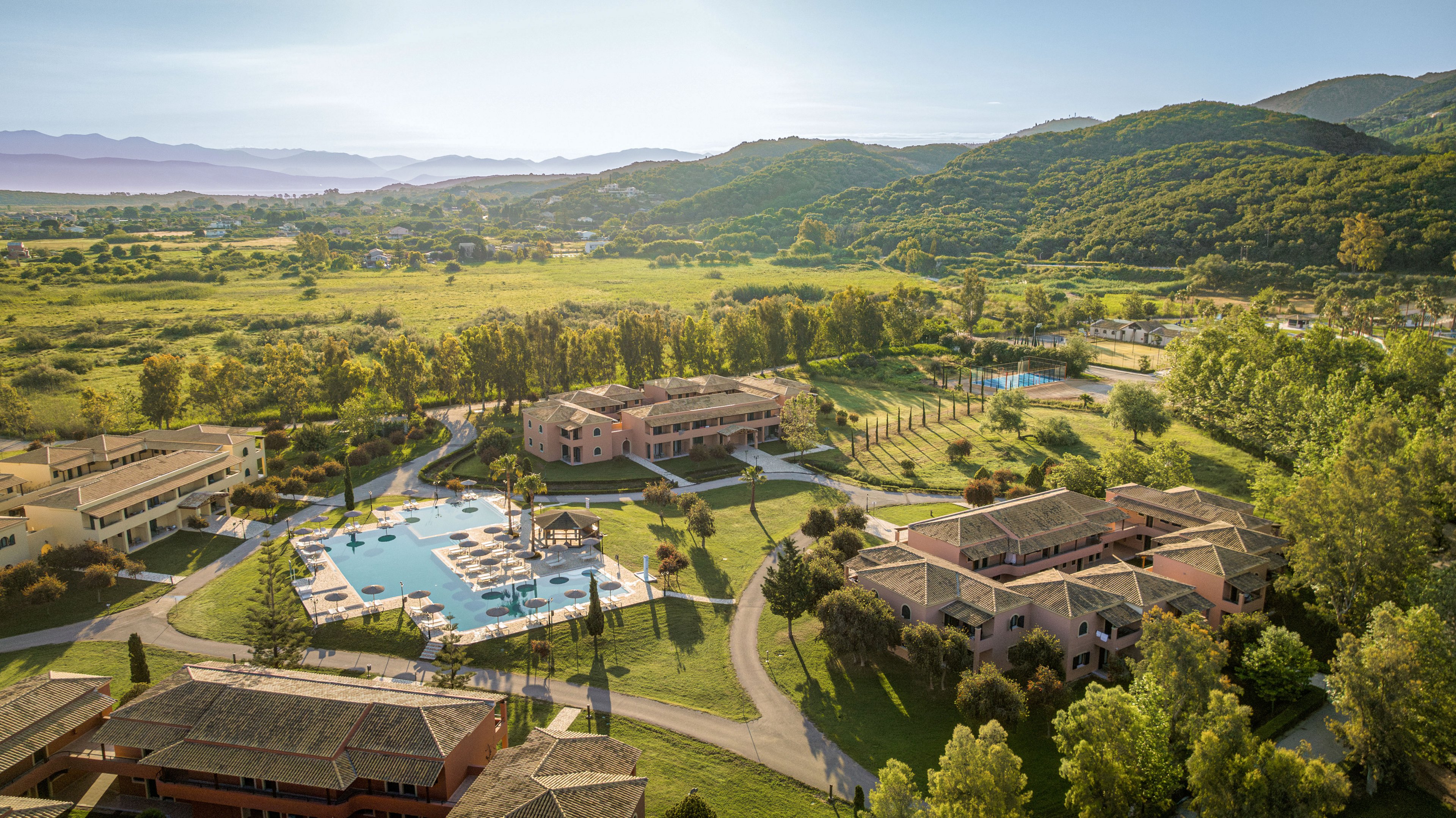 A large holiday resort complex is seen from above, featuring multiple low-rise buildings surrounding a central outdoor swimming pool. The property is set in a wide green valley with rolling forested hills in the background under a clear sky. Sun loungers and umbrellas line the pool area, and pathways connect the various structures and landscaped gardens. The overall scene suggests a tranquil vacation destination immersed in nature.