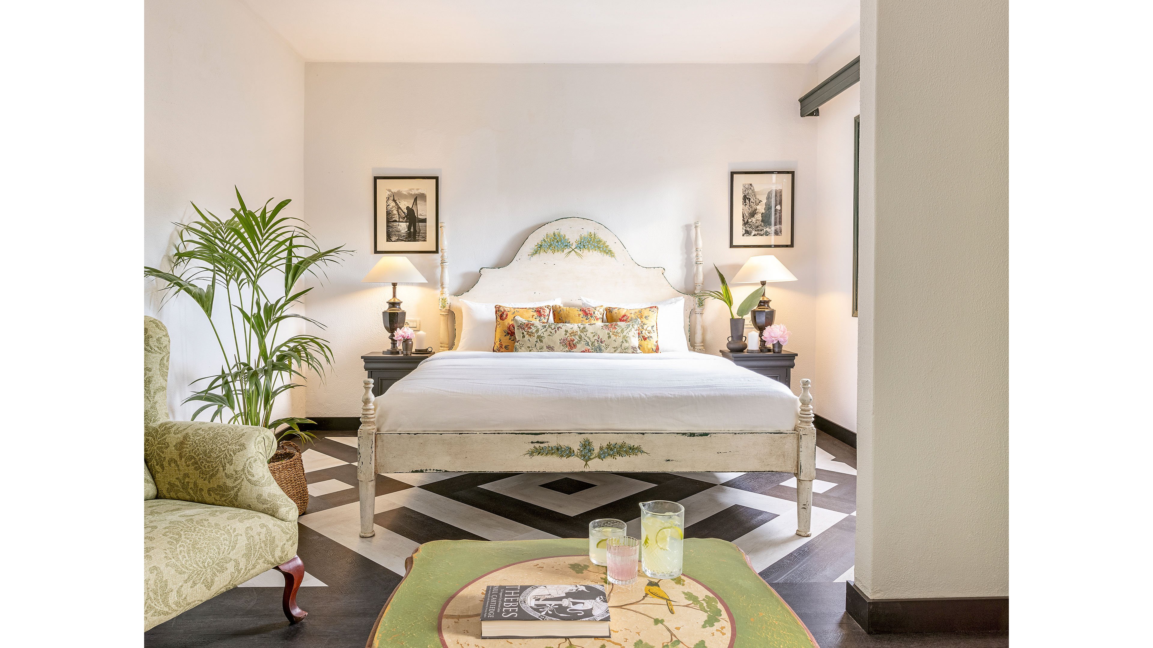 A light-filled bedroom features a vintage-style bed with a white headboard and floral accent pillows. Two framed artworks hang symmetrically above matching bedside tables with lamps and small decorative items. A patterned armchair and a round table with drinks and a book add cozy touches to the space. The room is finished with chevron floor tiles, potted plants, and soft neutral walls creating an inviting atmosphere.