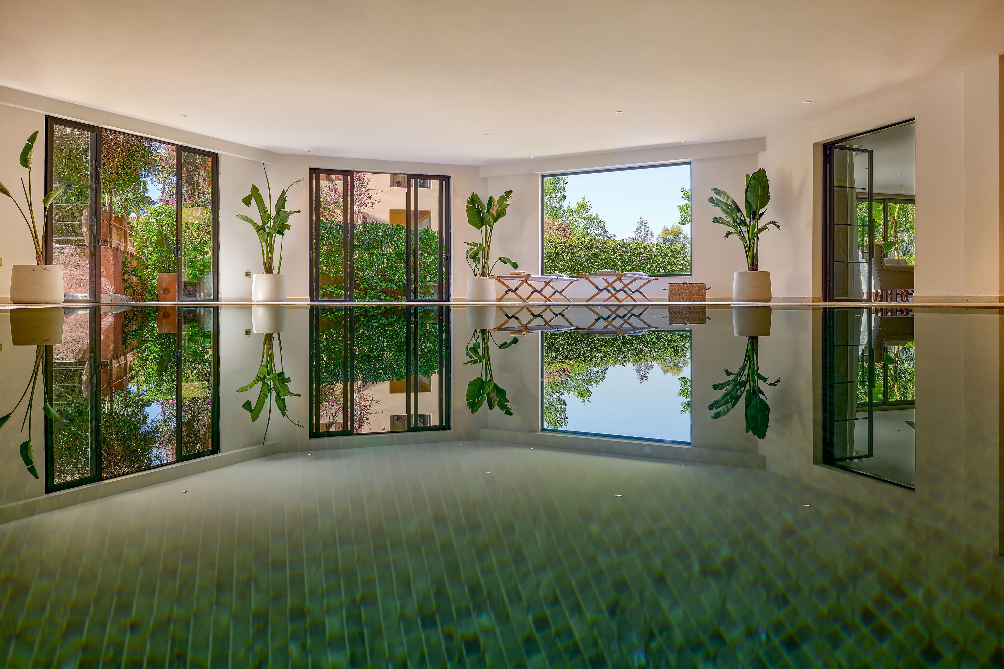 A serene indoor swimming pool reflects the surrounding architecture and greenery. Large floor-to-ceiling windows open onto a lush garden, filling the space with natural light. Potted tropical plants and a modern bench enhance the contemporary, minimalist design. The calm water surface and soft colors create a relaxing, spa-like atmosphere.