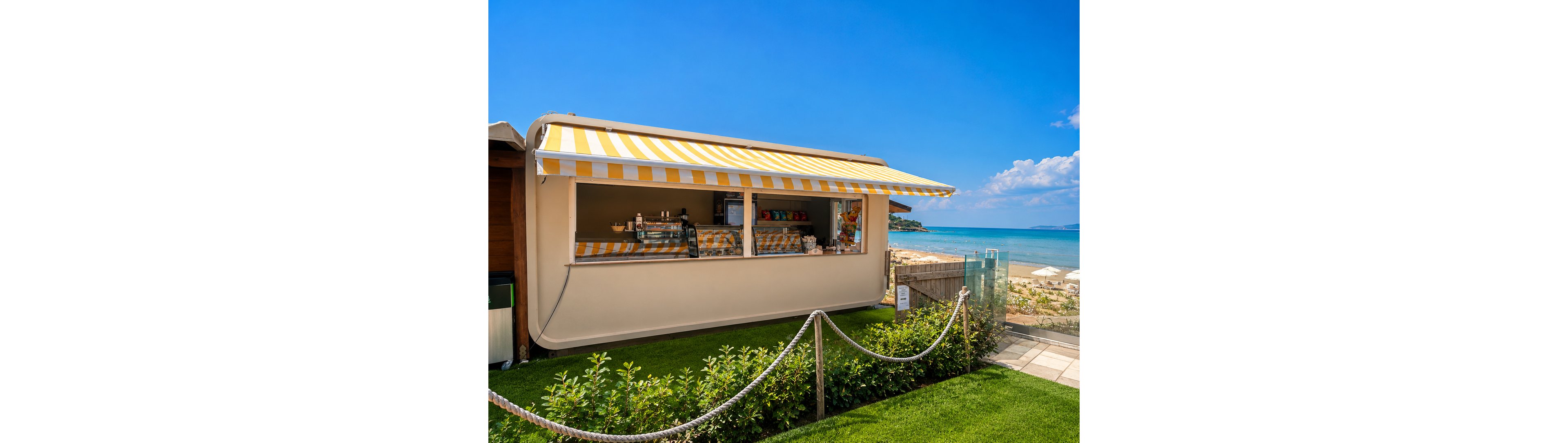 A small beachfront snack bar with a yellow and white striped awning stands on a neatly manicured lawn. The open service window reveals drinks and snacks arranged on the counter inside. A rope fence lines the foreground, leading toward a sandy beach with sun loungers and umbrellas. In the background, calm blue sea and a clear sky create a bright, summery coastal atmosphere.