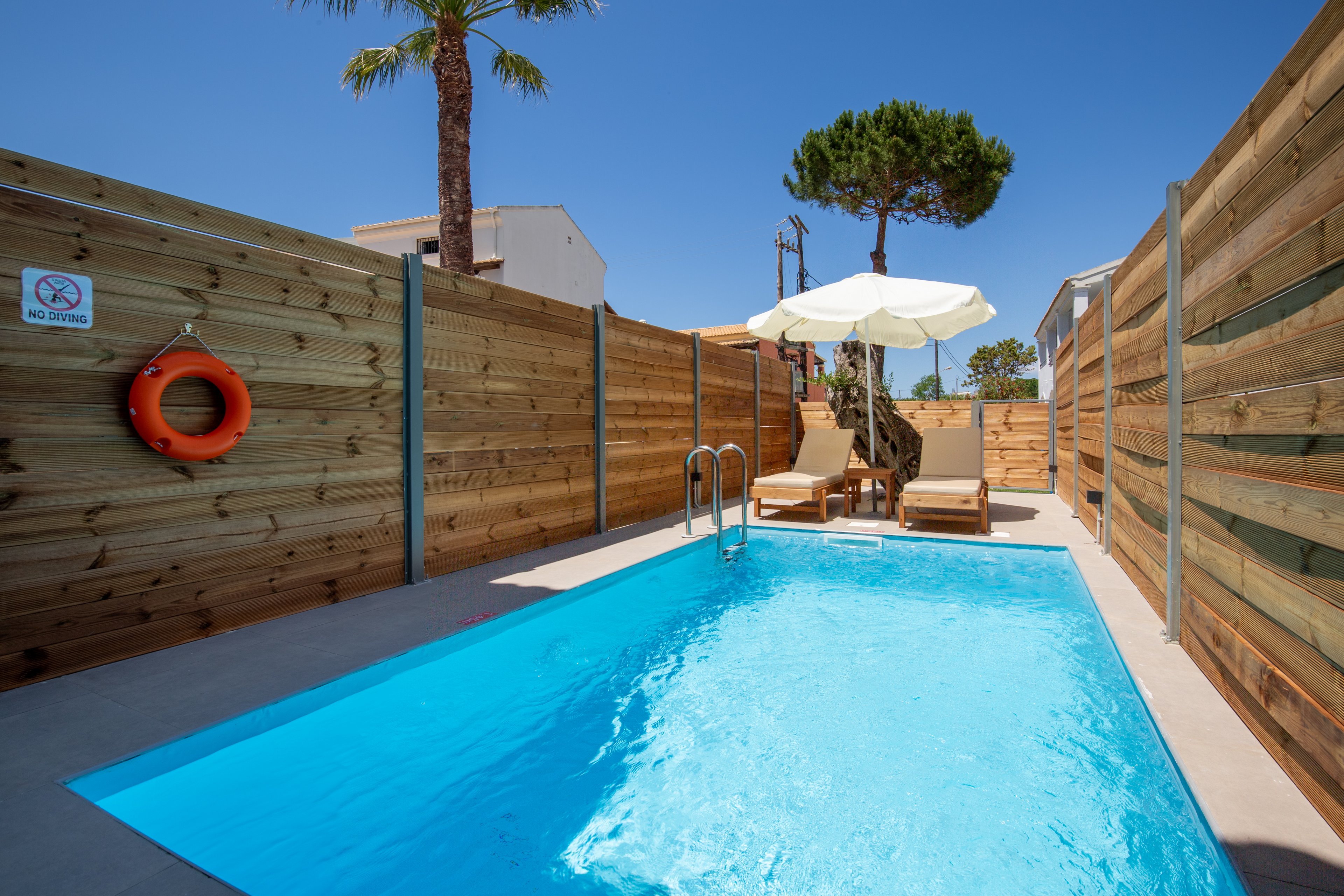 A small rectangular swimming pool is set in a private outdoor area surrounded by tall wooden fencing. At the far end, two sun loungers sit under a large white umbrella, creating a relaxing spot for sunbathing. A bright orange lifebuoy hangs on the left fence near a safety sign, and a metal ladder provides access to the clear blue water. In the background, tall trees and a clear blue sky suggest a warm, sunny day.