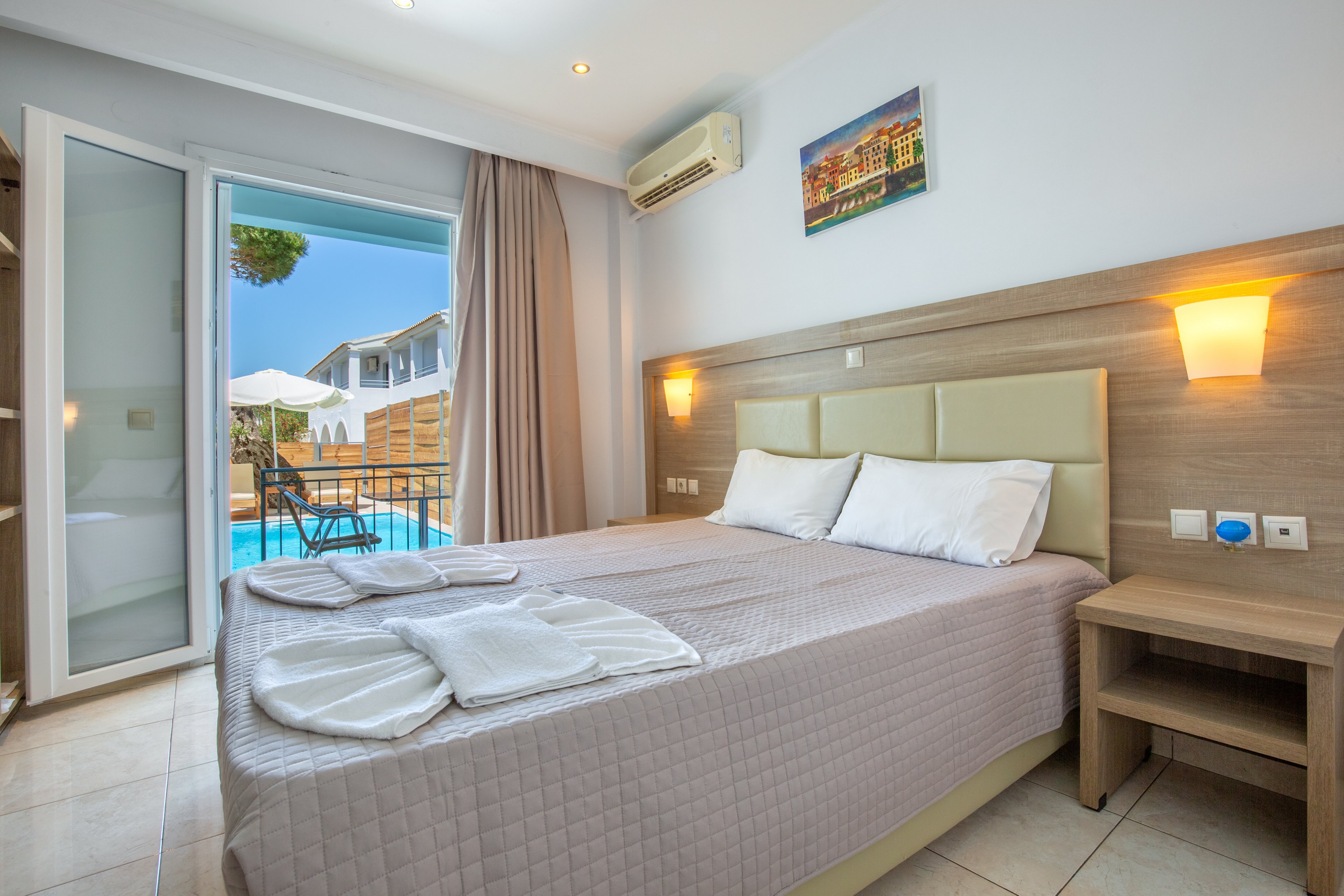 A neatly made double bed with folded white towels is the focal point of a bright hotel bedroom. The room features light wood furniture, wall-mounted bedside lamps, and an air conditioning unit above the bed. An open glass door leads to a balcony overlooking a swimming pool and sun loungers outside. Soft neutral colors and natural daylight create a clean, inviting atmosphere suitable for vacation or short stays.