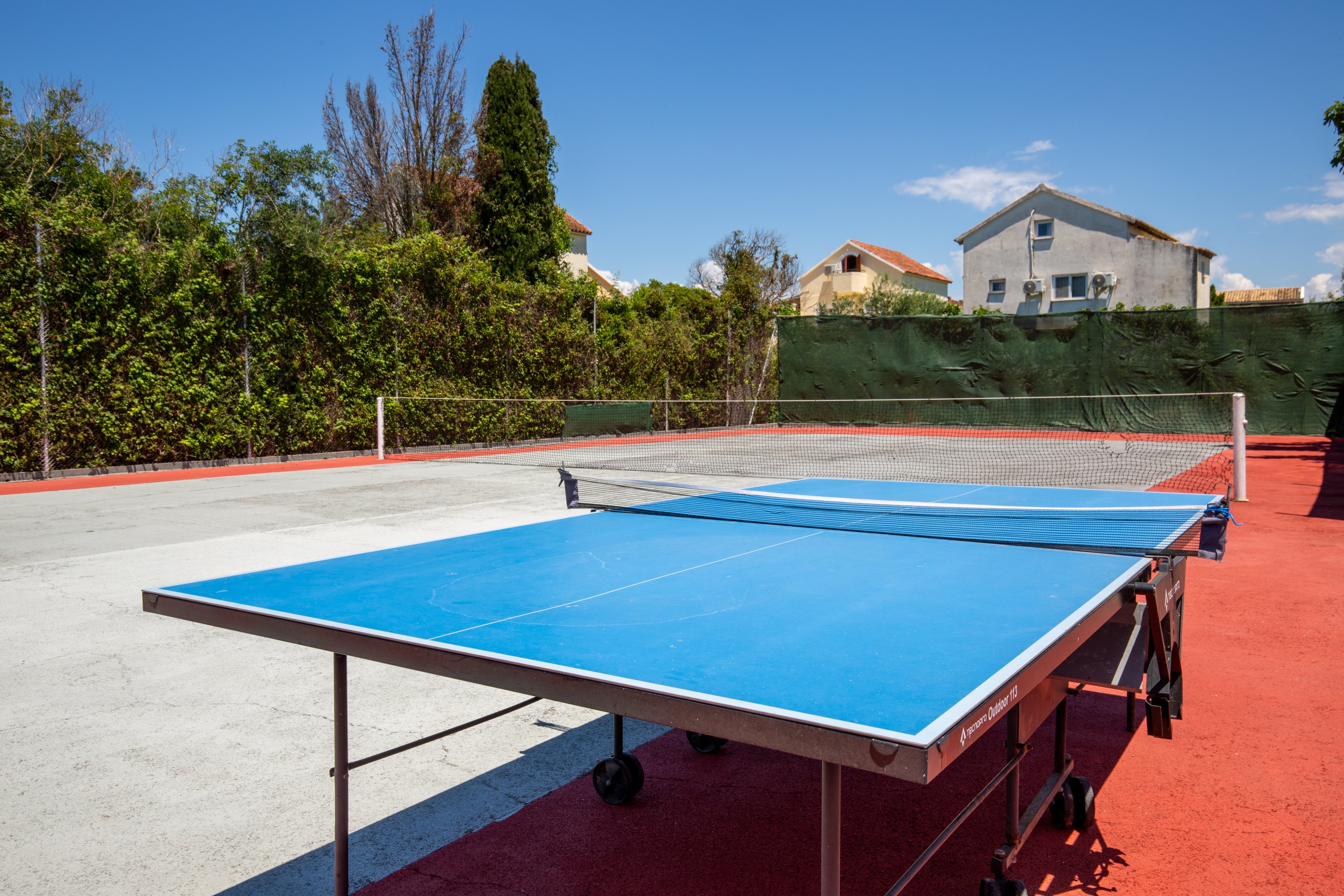 A blue table tennis table stands on an outdoor sports court under a clear blue sky. The court surface combines red and light gray areas, with a tennis net visible in the background. Tall green hedges and residential houses surround the court, creating a private recreational setting. Bright daylight and vivid colors give the scene a clean, summery atmosphere.