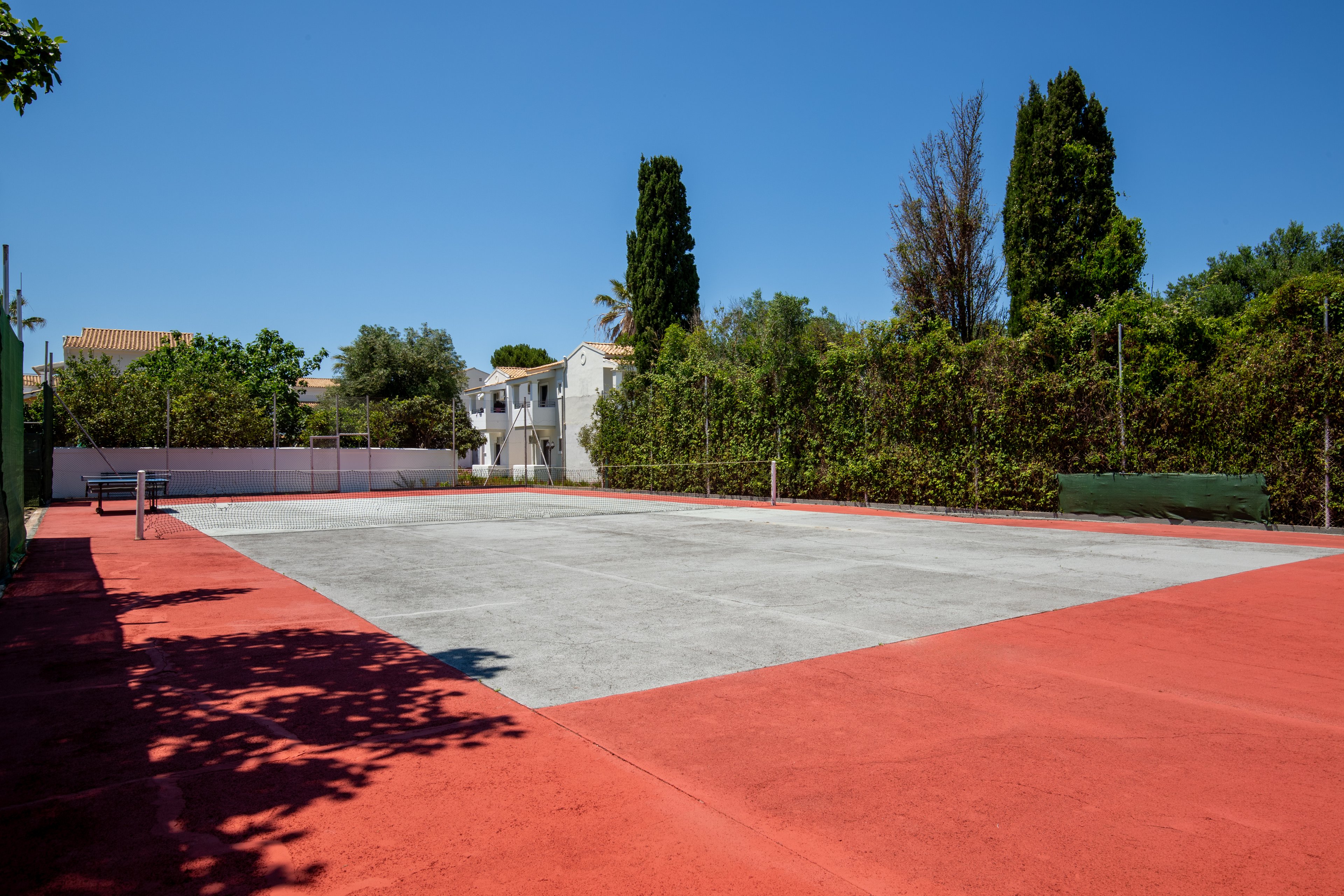 An empty outdoor tennis court sits under a clear blue sky in a residential area. The court surface is light gray with a red border and surrounded by tall green trees and hedges. In the background, white houses with balconies and tiled roofs are visible. The bright daylight emphasizes the clean, quiet, and well-maintained sports setting.