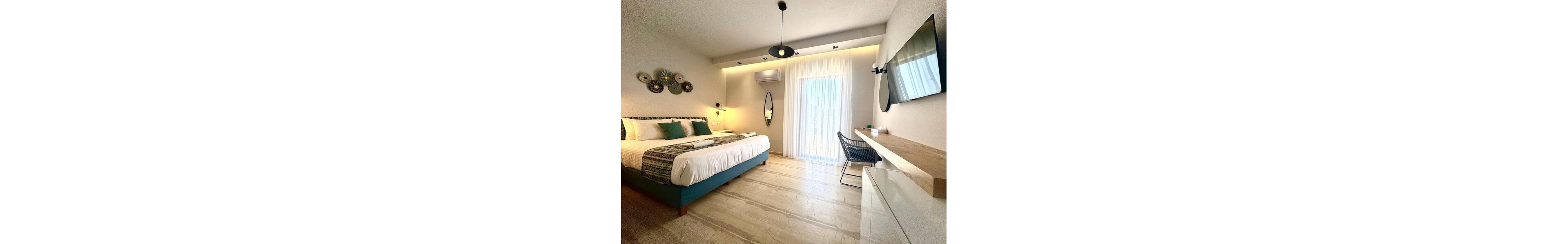 Bright contemporary bedroom features a large double bed with teal base and coordinated cushions. The room includes a wall-mounted television, floating wooden desk, and a woven chair, creating a clean minimalist workspace. Sheer curtains cover a glass door leading to a balcony, allowing soft natural light to fill the space. Decorative wall art, an oval mirror, and warm recessed lighting add a cozy, inviting atmosphere.