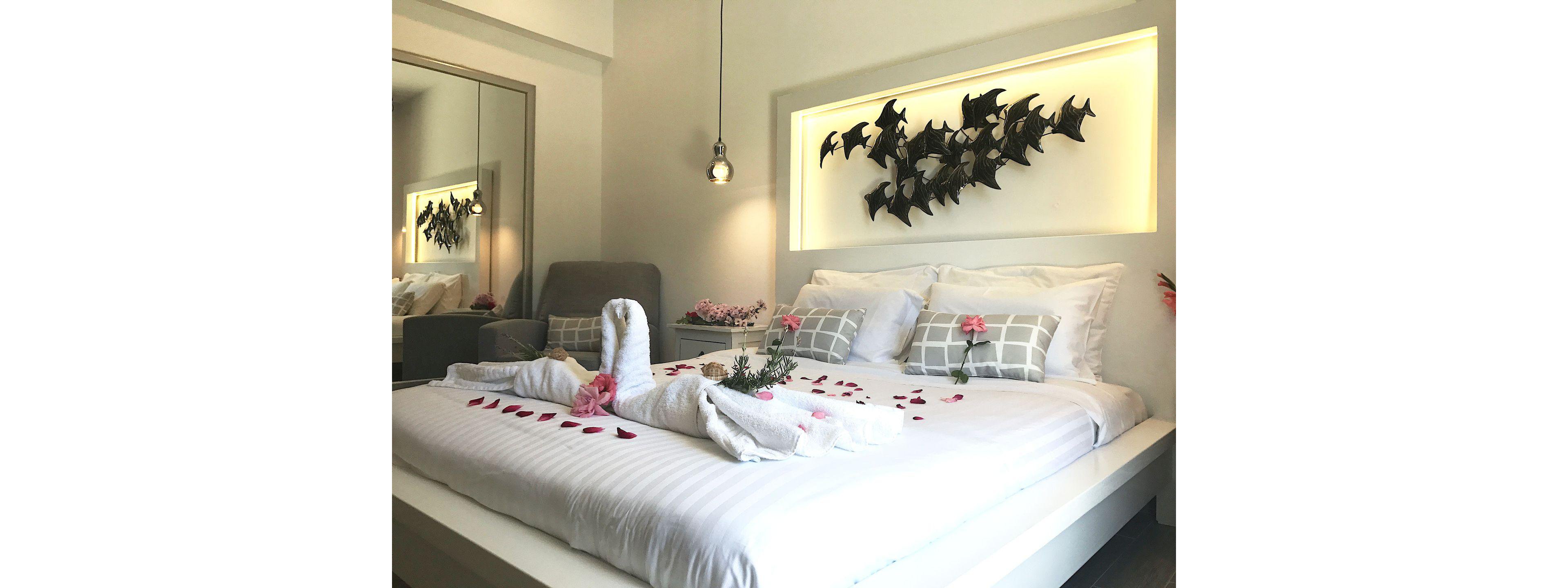 A modern hotel bedroom features a neatly made bed with white linens and decorative gray plaid pillows. A towel folded into a swan shape is placed at the center of the bed, surrounded by pink flowers and scattered rose petals. Above the bed, a lit recessed wall niche displays a sculptural art piece of multiple black birds in flight. A hanging pendant light, upholstered chair, and mirrored wardrobe complete the contemporary, romantic setting.