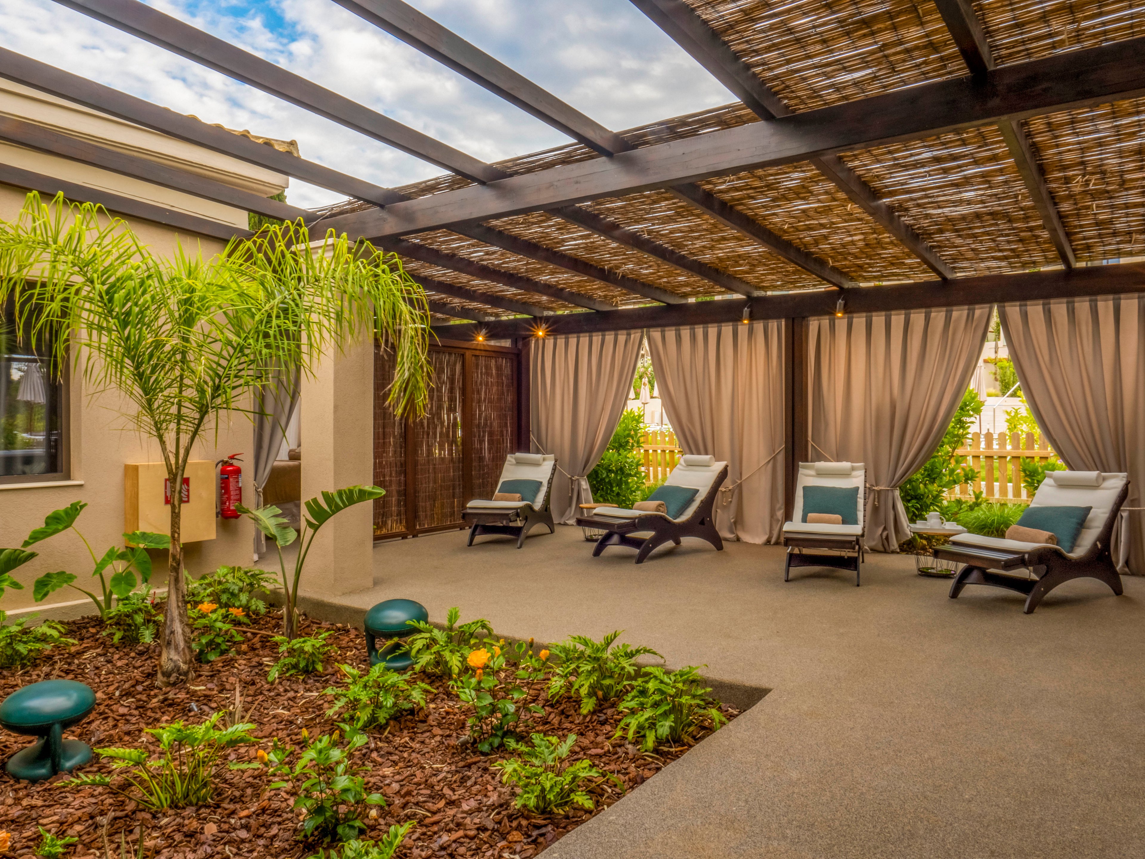 An inviting outdoor spa lounge features several cushioned recliners arranged beneath a wooden pergola. Beige curtains hang between the posts, creating a sense of privacy and shade. A small landscaped garden with tropical plants and mulch runs along the center of the space. Soft natural light and neutral tones contribute to a calm, relaxing atmosphere suitable for rest and wellness.