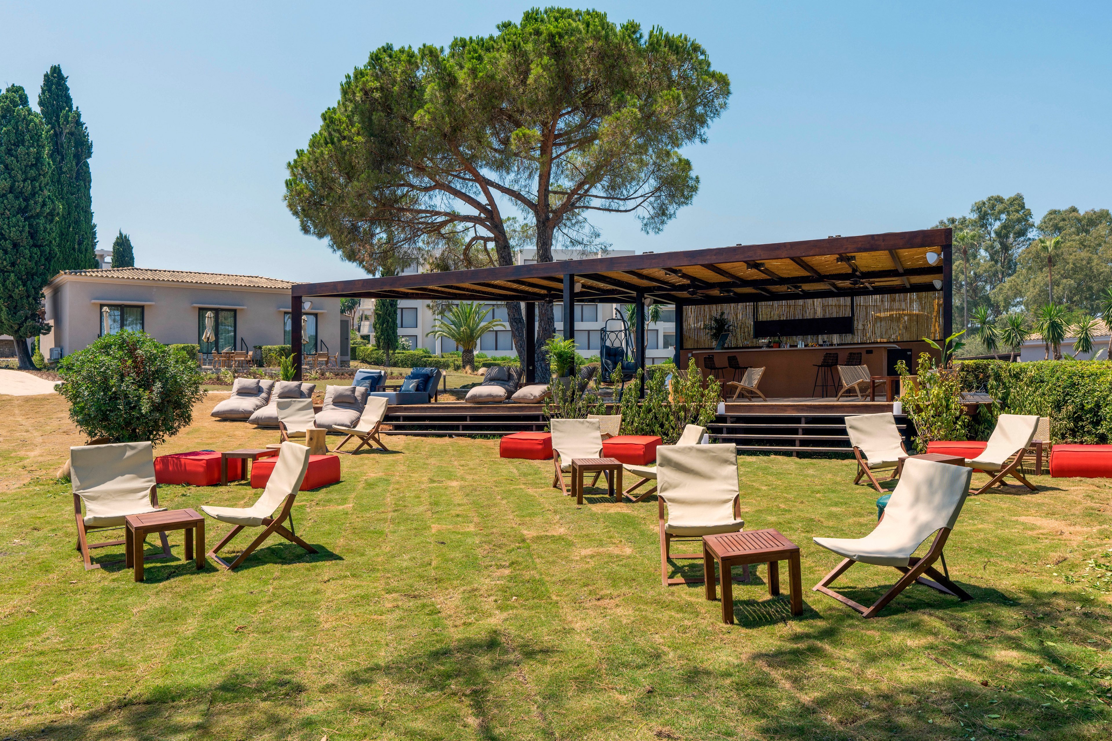 An open-air lounge area is set on a neatly mowed lawn with several canvas deck chairs and small wooden tables. Cushions and low seating are arranged near a modern wooden bar structure shaded by a pergola. A large tree provides additional shade, while surrounding greenery and distant trees frame the relaxed setting. The scene suggests a tranquil resort or garden venue on a sunny day.
