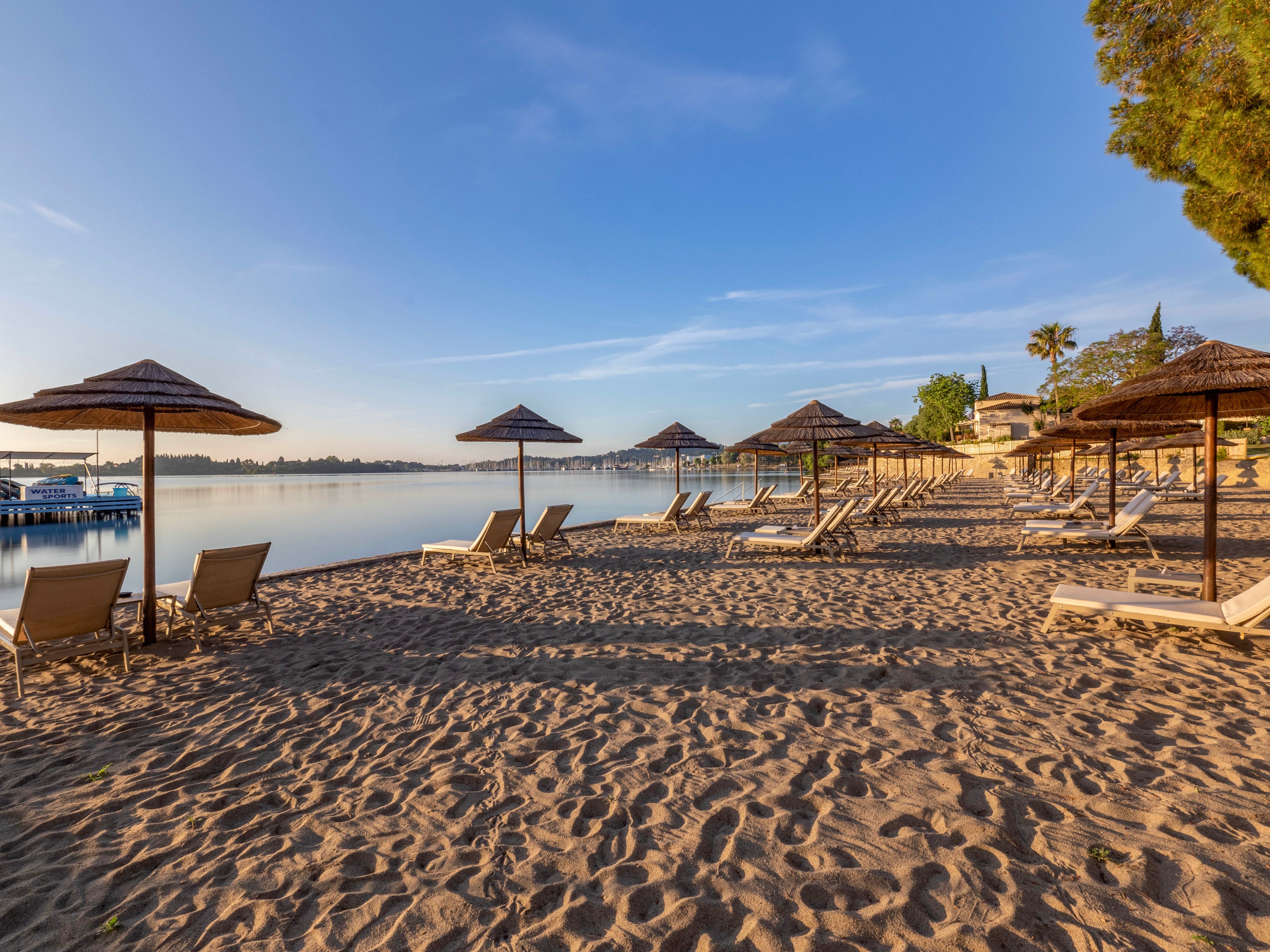 A tranquil sandy beach is lined with empty sun loungers and straw parasols facing a calm body of water. The early morning light casts long shadows across the sand, creating a peaceful and inviting atmosphere. Buildings, trees, and a small pier are visible in the background along the shoreline. The clear blue sky and still water emphasize the serene coastal setting.