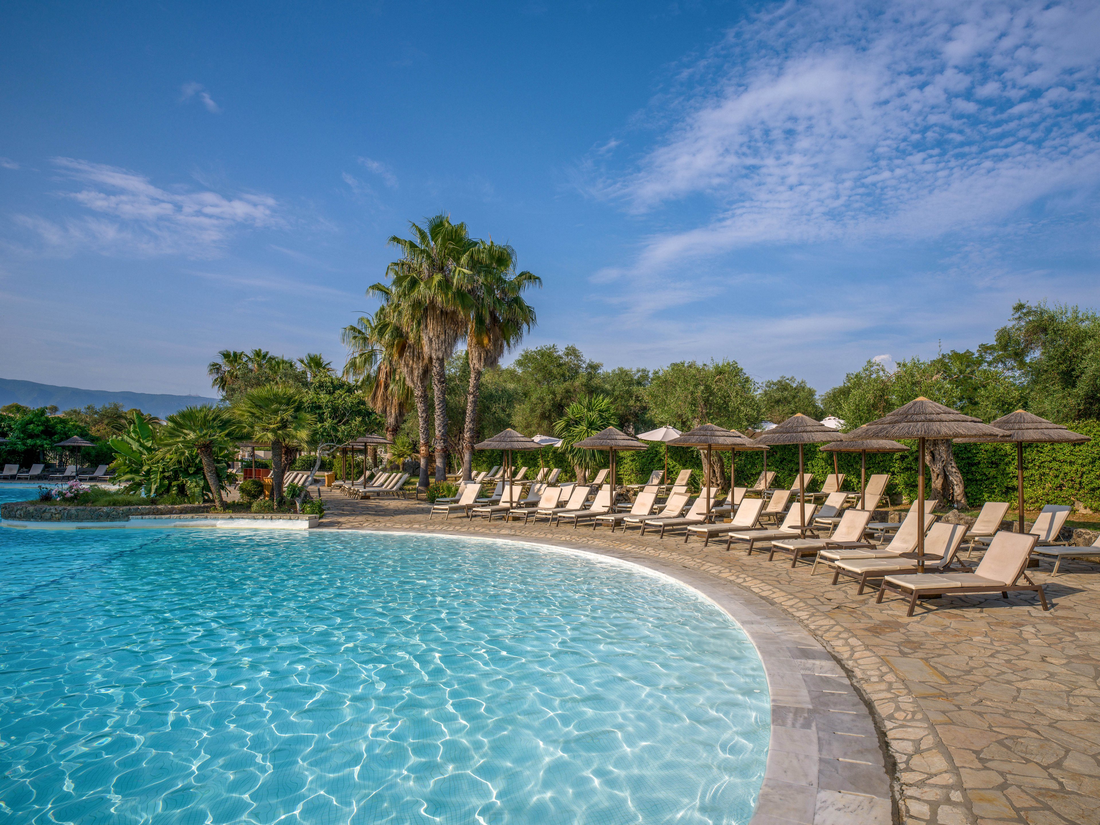 A large curved swimming pool sparkles under bright daylight at a resort. Numerous empty sun loungers with straw parasols line the stone poolside terrace. Tall palm trees and lush greenery surround the area, with distant hills visible under a blue sky. The tranquil scene suggests a relaxing vacation atmosphere in warm weather.