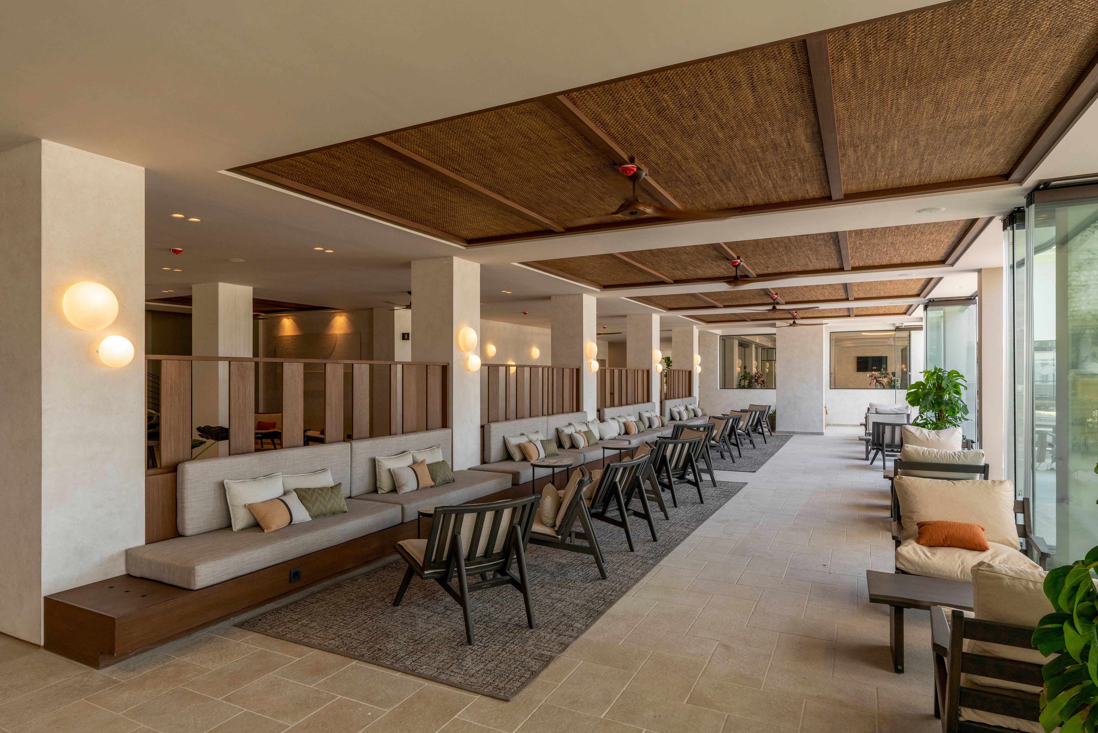 Spacious contemporary lounge area with long cushioned benches and individual chairs arranged in rows. The setting appears to be an open-air or semi-outdoor hotel or resort space with large glass walls on one side. Warm neutral tones, wooden ceiling panels, and soft lighting create a relaxed, upscale atmosphere. Potted plants and accent pillows add comfort and a touch of greenery to the minimalist design.