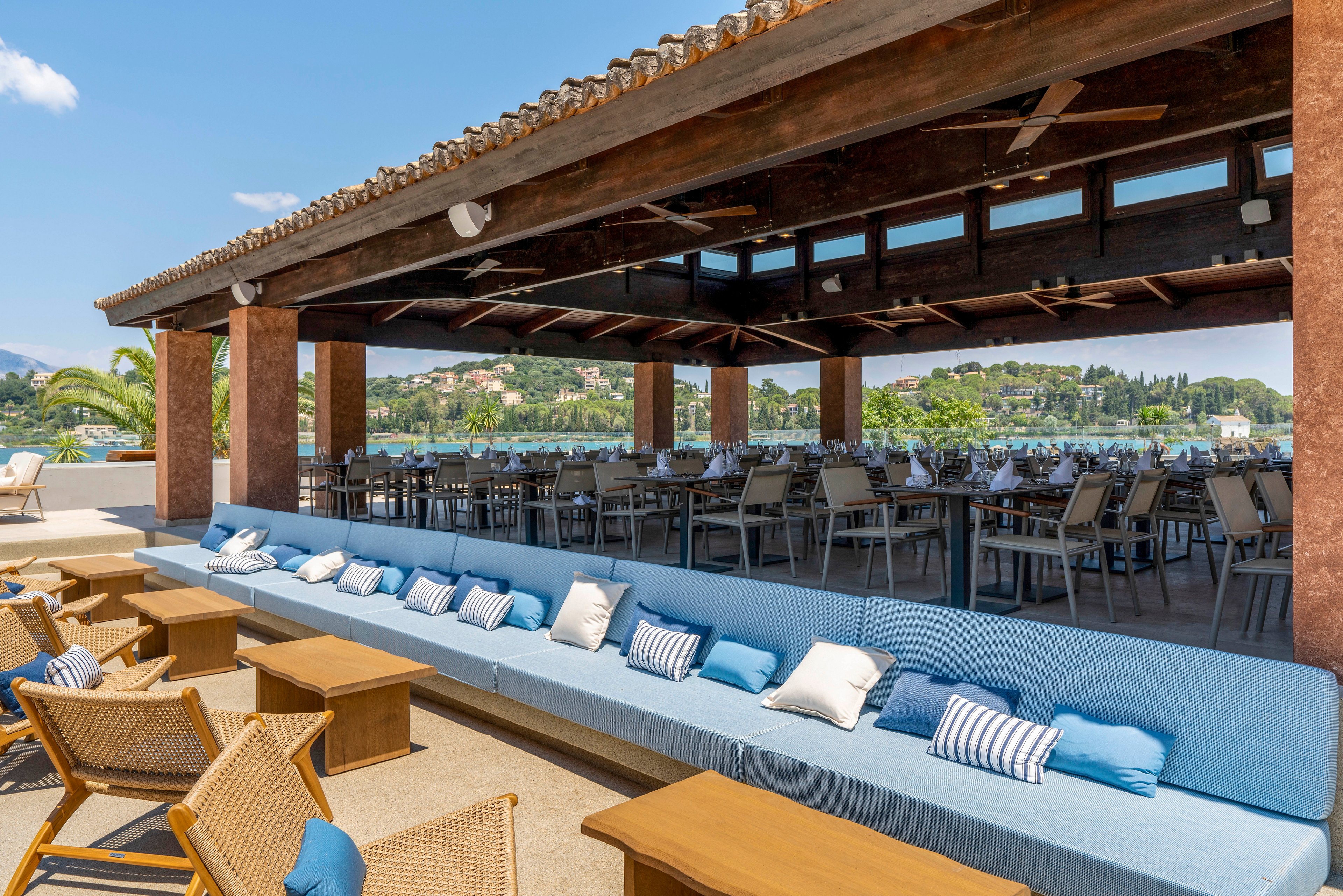 An open-air restaurant with a wooden roof overlooks a scenic lakeside or riverside view. Long blue cushioned benches with assorted throw pillows line the foreground, facing wicker lounge chairs and low wooden tables. In the background, rows of dining tables and chairs are neatly arranged under the shaded pavilion. The bright daylight and clear sky create a relaxed, resort-like atmosphere.