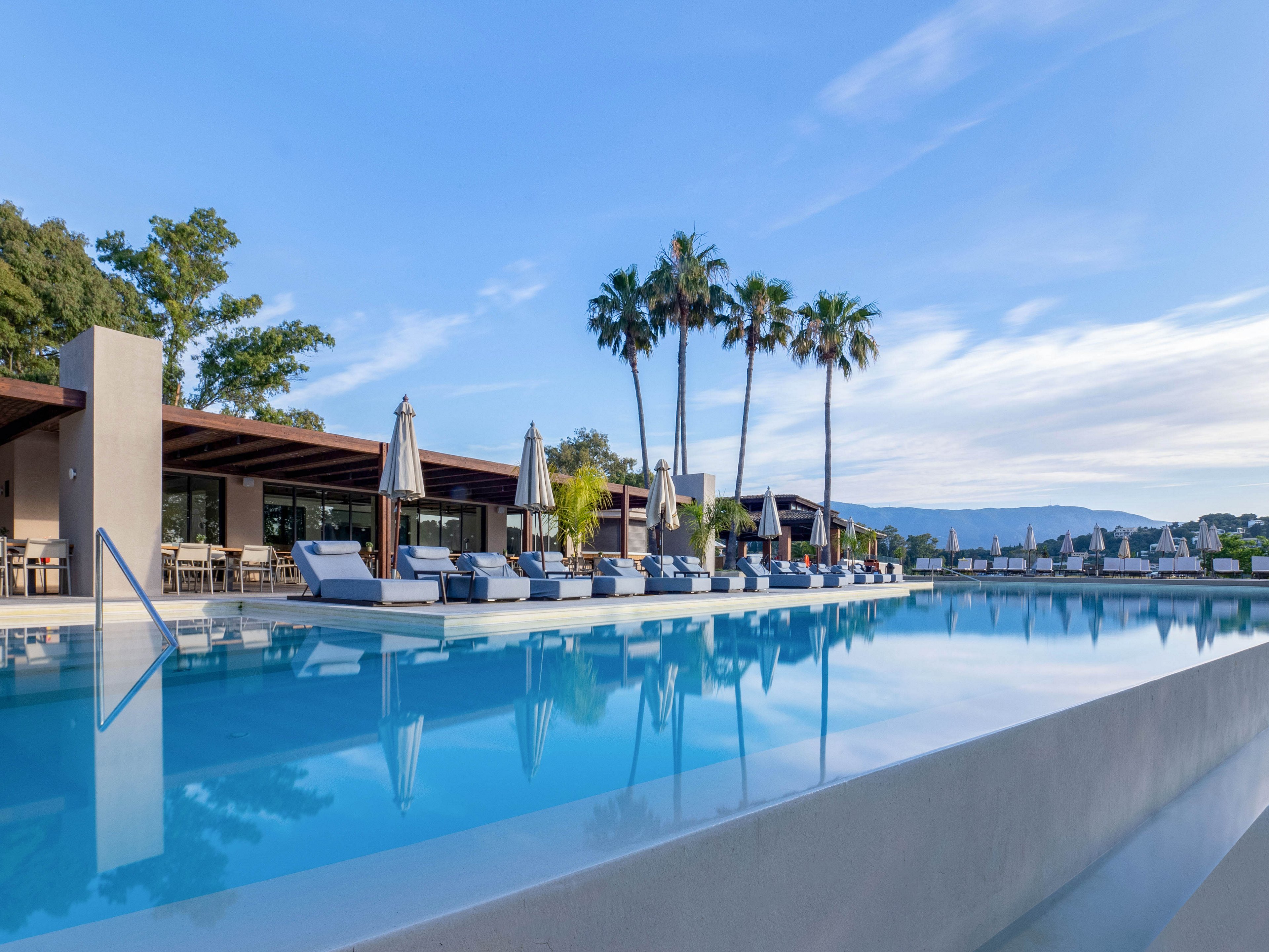 A large, modern swimming pool stretches alongside a contemporary resort building. Sun loungers and closed umbrellas line the pool deck, suggesting a quiet, early or late day setting. Tall palm trees rise behind the pool, with distant hills and a clear blue sky in the background. The calm water reflects the surrounding architecture and trees, creating a serene vacation atmosphere.