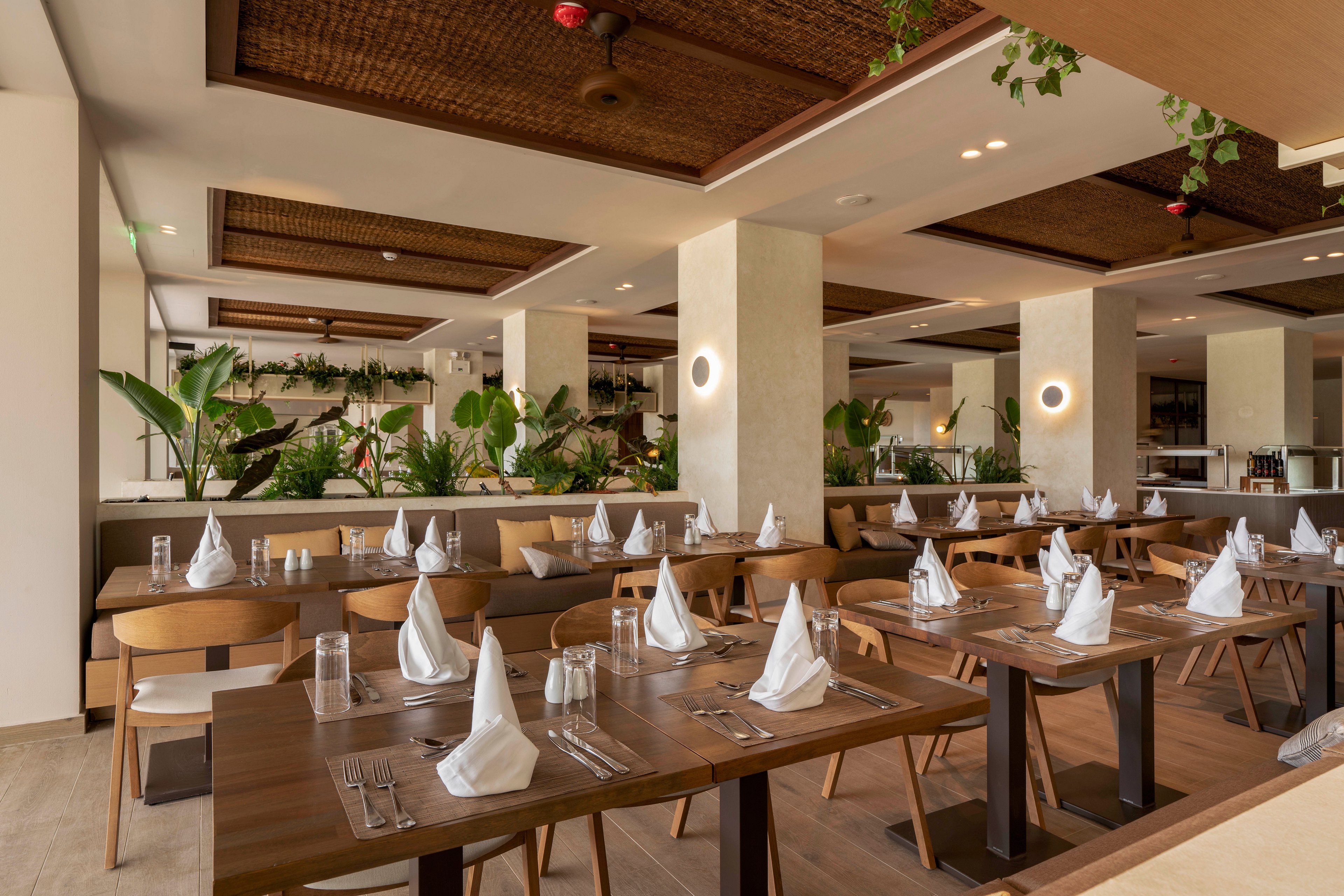A contemporary restaurant dining area features neatly arranged wooden tables set with white napkins and clear glassware. The space is bright and spacious, with neutral walls, light flooring, and large pillars dividing the room. Green plants and foliage add a natural accent along the perimeter and above some partitions. Warm ceiling lighting and wall sconces create a welcoming, relaxed atmosphere in the empty dining room.