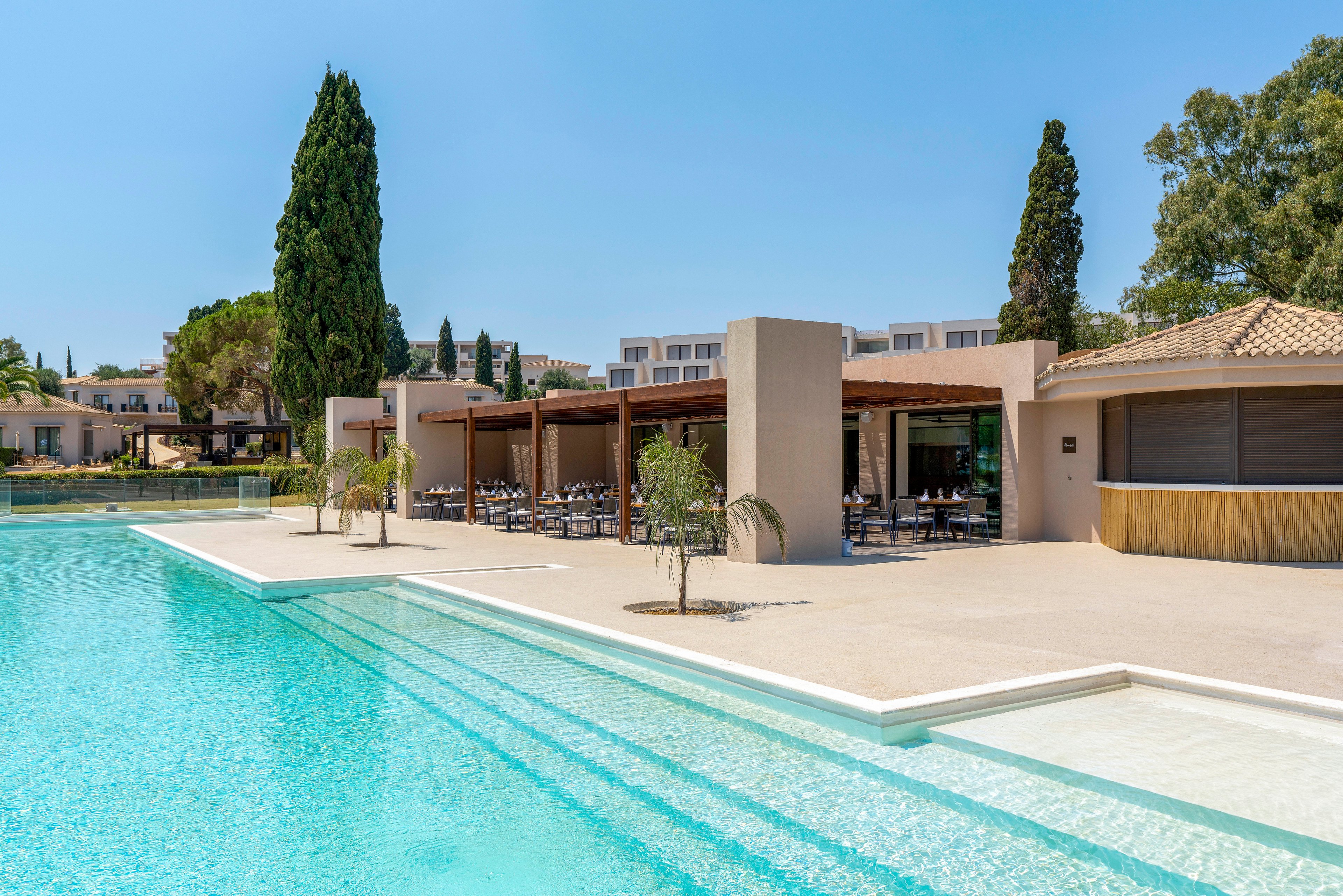 A large turquoise swimming pool stretches alongside a modern resort terrace. The open-air bar and dining area sit under a flat roof with clean architectural lines. Tall cypress trees and other greenery frame the scene under a clear blue sky. The bright sunlight and minimal design create a calm, luxurious vacation atmosphere.