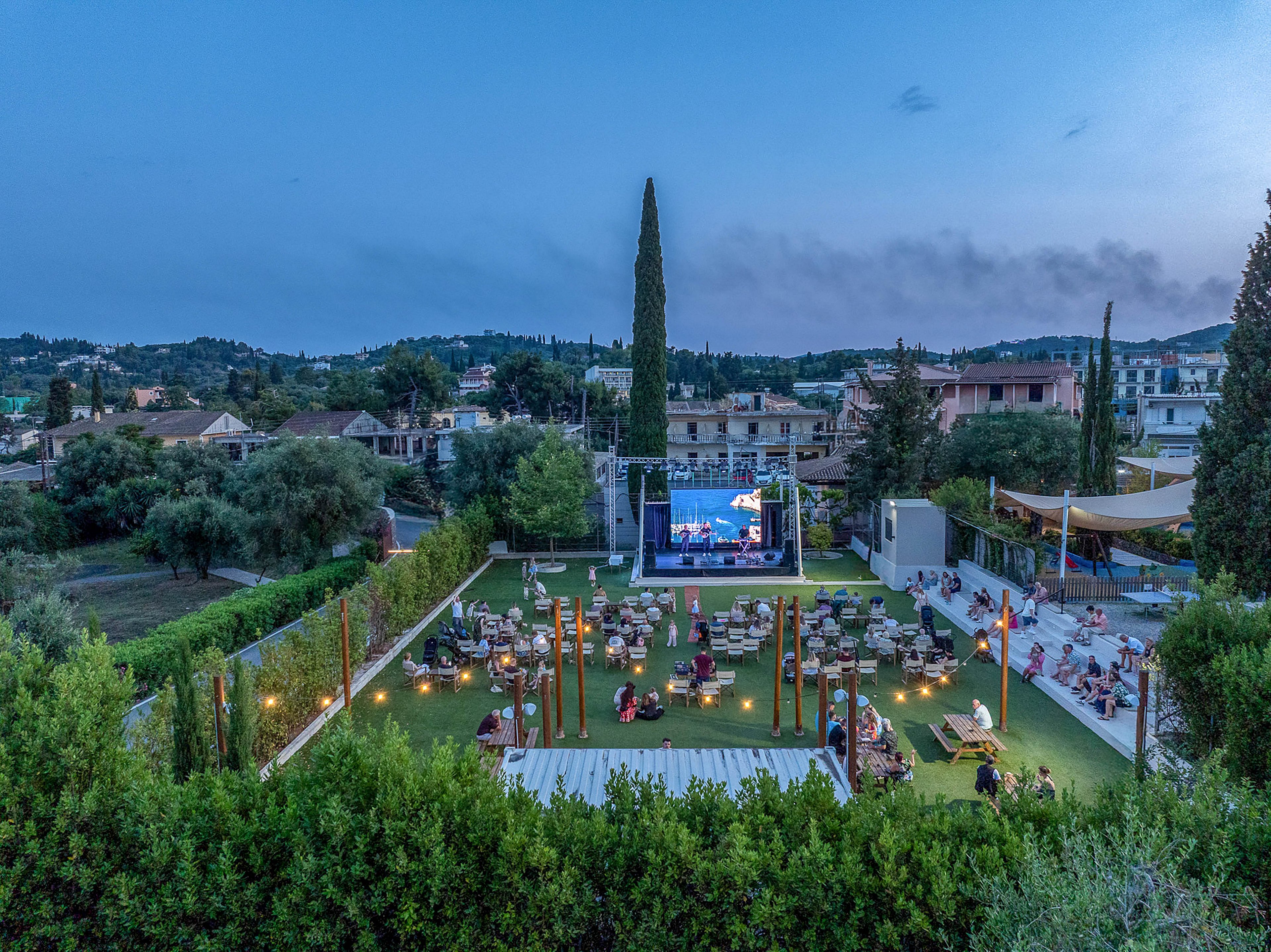 An open-air cinema is set up in a landscaped garden with rows of tables and chairs facing a large screen. People are seated and walking around as evening falls, with warm lights illuminating the lawn. The venue is surrounded by tall hedges, trees, and a mix of residential buildings in the background. The sky is a soft blue, adding a relaxed summer atmosphere to the scene.