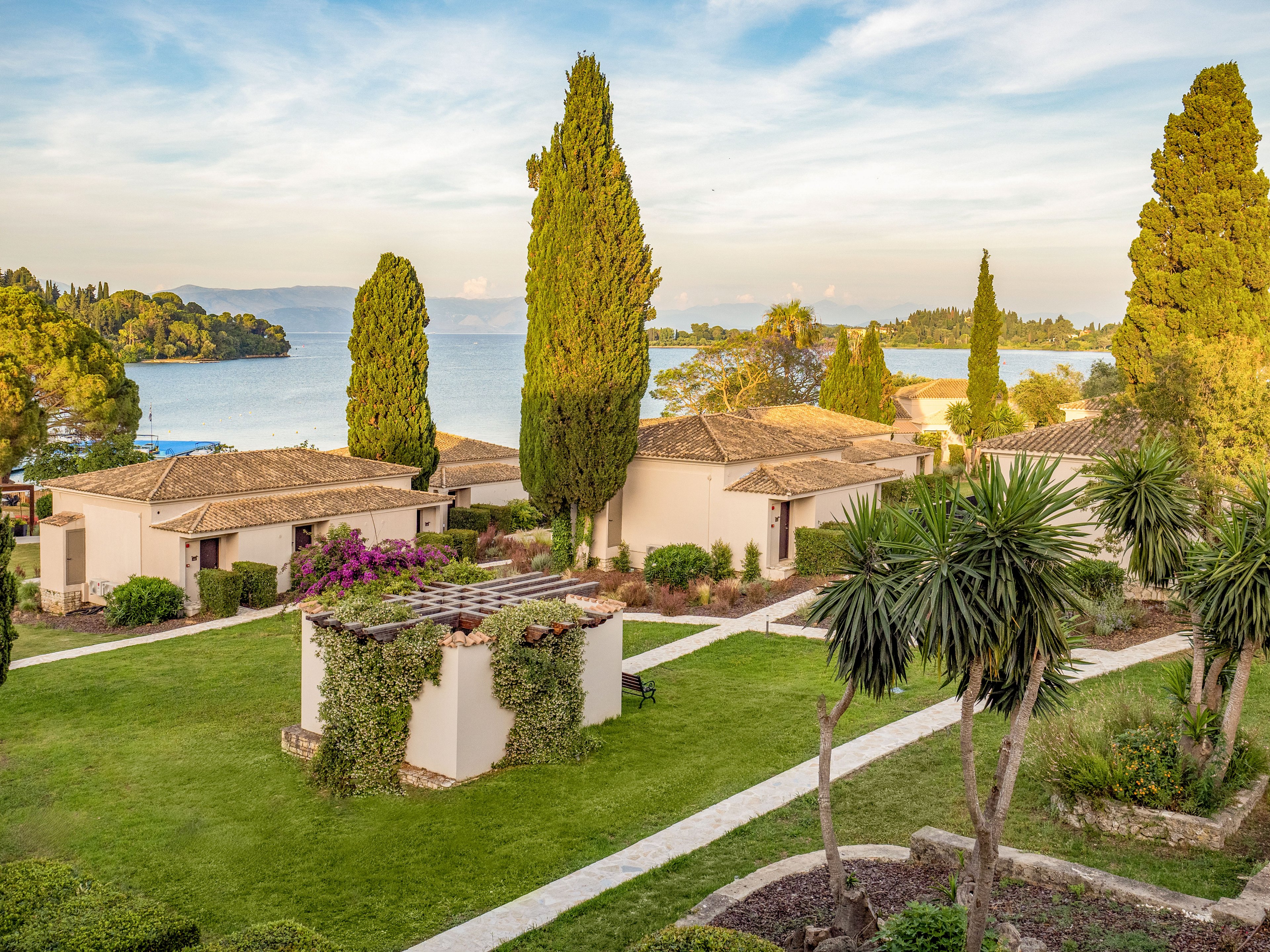 A cluster of light-colored villas sits within a manicured coastal resort overlooking calm blue water. Tall cypress trees, palm-like plants, and flowering shrubs line the pathways between the buildings. The scene is bathed in warm, late-afternoon sunlight, creating soft shadows and a tranquil atmosphere. In the distance, tree-covered hills and a serene bay complete the picturesque Mediterranean landscape.