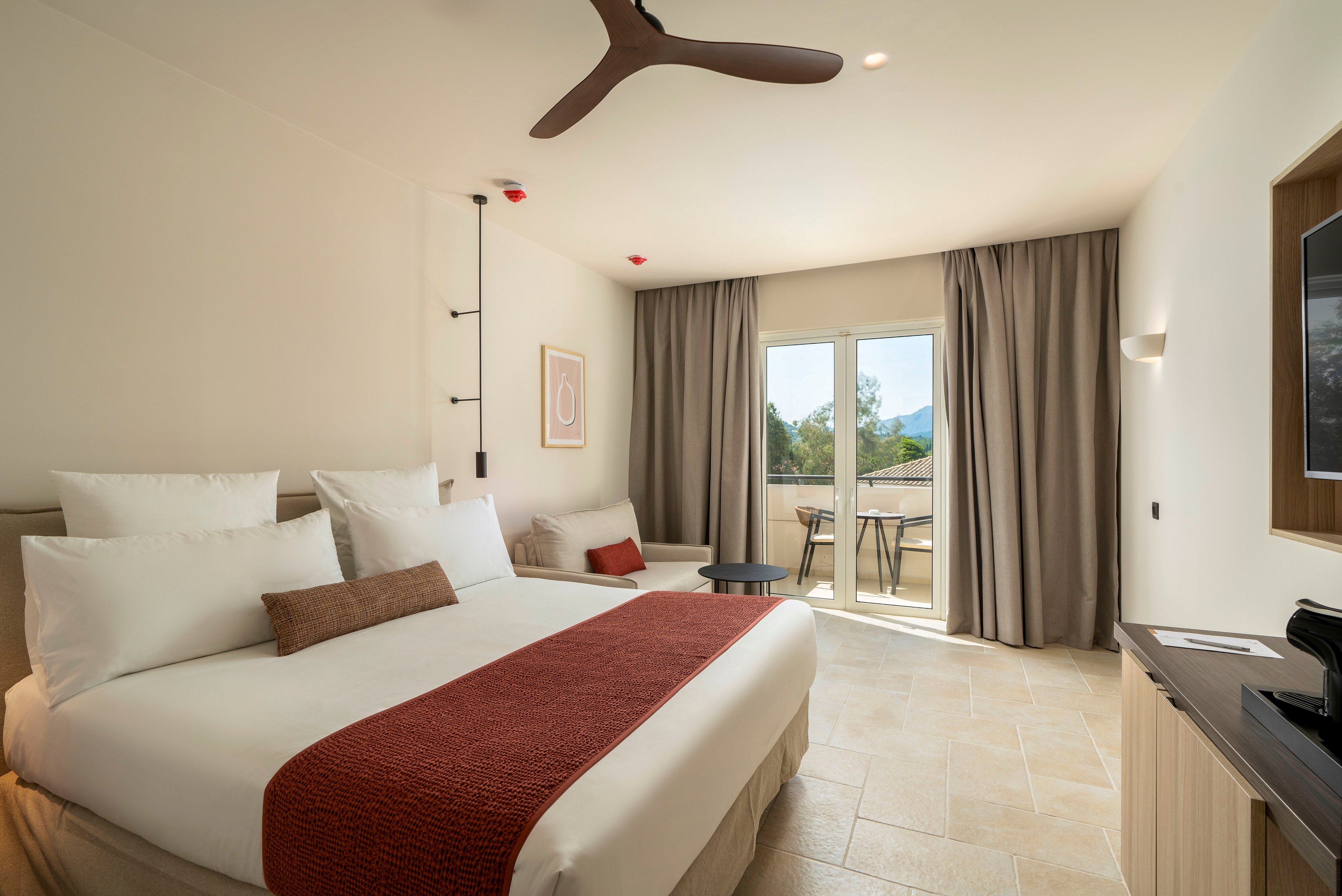 A contemporary hotel bedroom features a large neatly made bed with white linens and a rust-colored throw. Neutral walls, tiled flooring, and minimalist artwork create a calm, modern atmosphere. Floor-to-ceiling curtains frame glass doors that open onto a terrace with outdoor seating and a scenic view of trees and distant hills. A ceiling fan, wall-mounted lighting, and a media console complete the comfortable guest room setting.