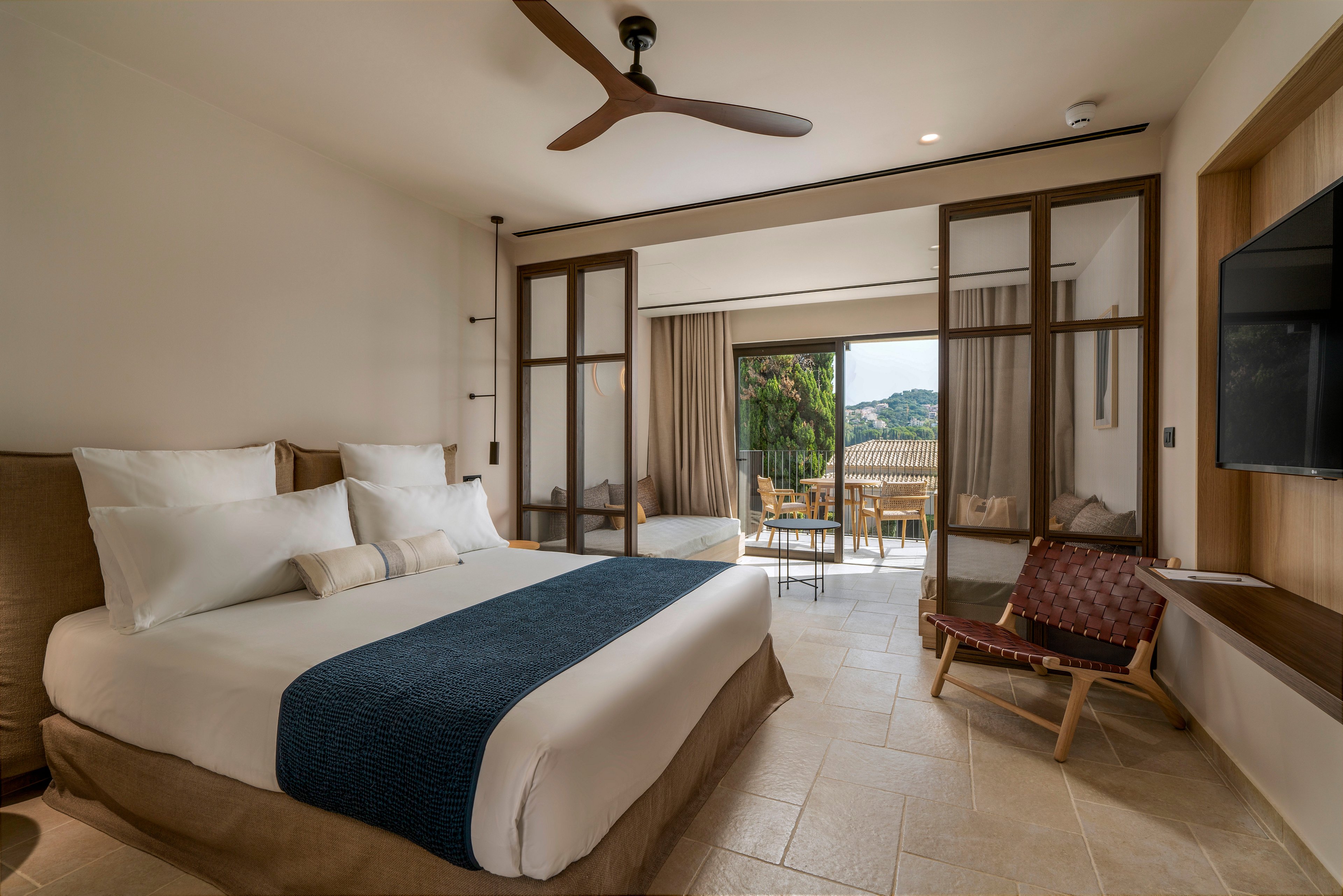 A contemporary hotel bedroom features a large bed with white linens and a dark blue throw blanket. The room opens onto a balcony through sliding glass doors, revealing an outdoor seating area and a scenic hillside view. Neutral tones, wooden accents, and a ceiling fan create a calm, minimalist atmosphere. A wall-mounted television and a woven lounge chair complete the cozy, modern interior.