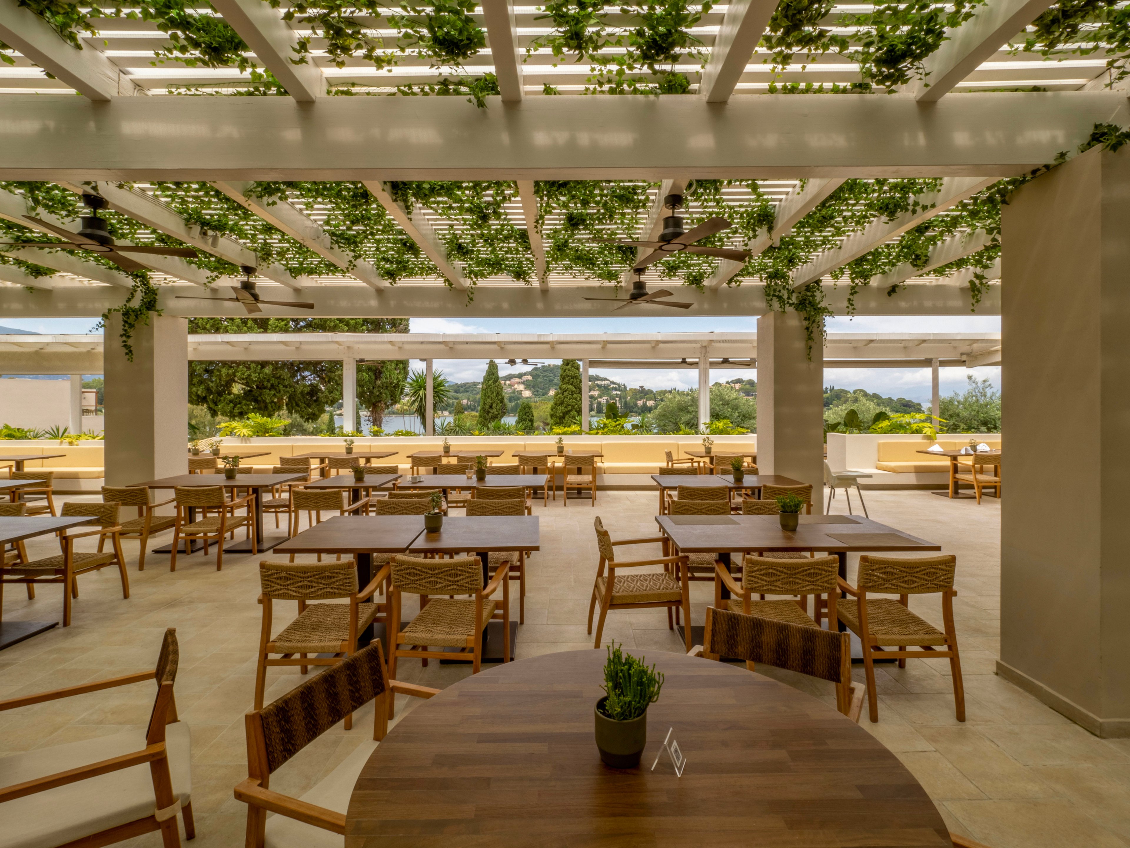 An open-air terrace restaurant features wooden tables and chairs arranged in neat rows. A pergola ceiling above is covered with green vines, creating a natural canopy and filtered light. The space overlooks a scenic landscape with trees and distant hills, giving the area a relaxed, vacation-like atmosphere. Small potted plants on the tables add a touch of greenery and complement the warm, neutral tones of the decor.